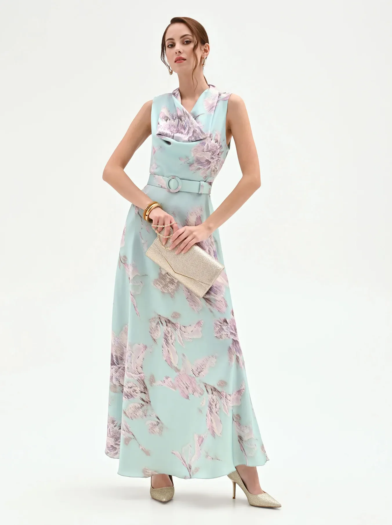 Woman in a pastel blue floral gown with a cowl neckline holds a textured clutch. She wears gold bracelets and beige heels, posing elegantly on a plain background.