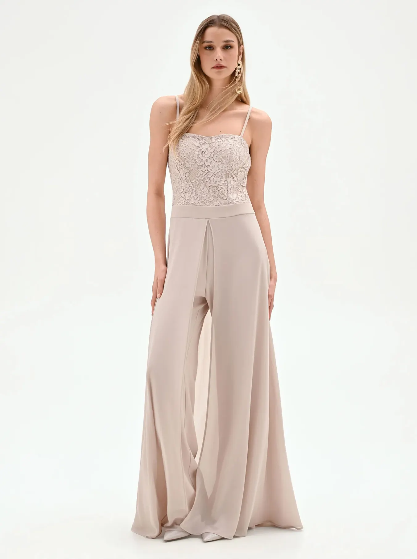 A woman stands confidently in a beige jumpsuit with a delicate lace bodice and flowing wide-leg pants. The neutral tones convey elegance and sophistication.