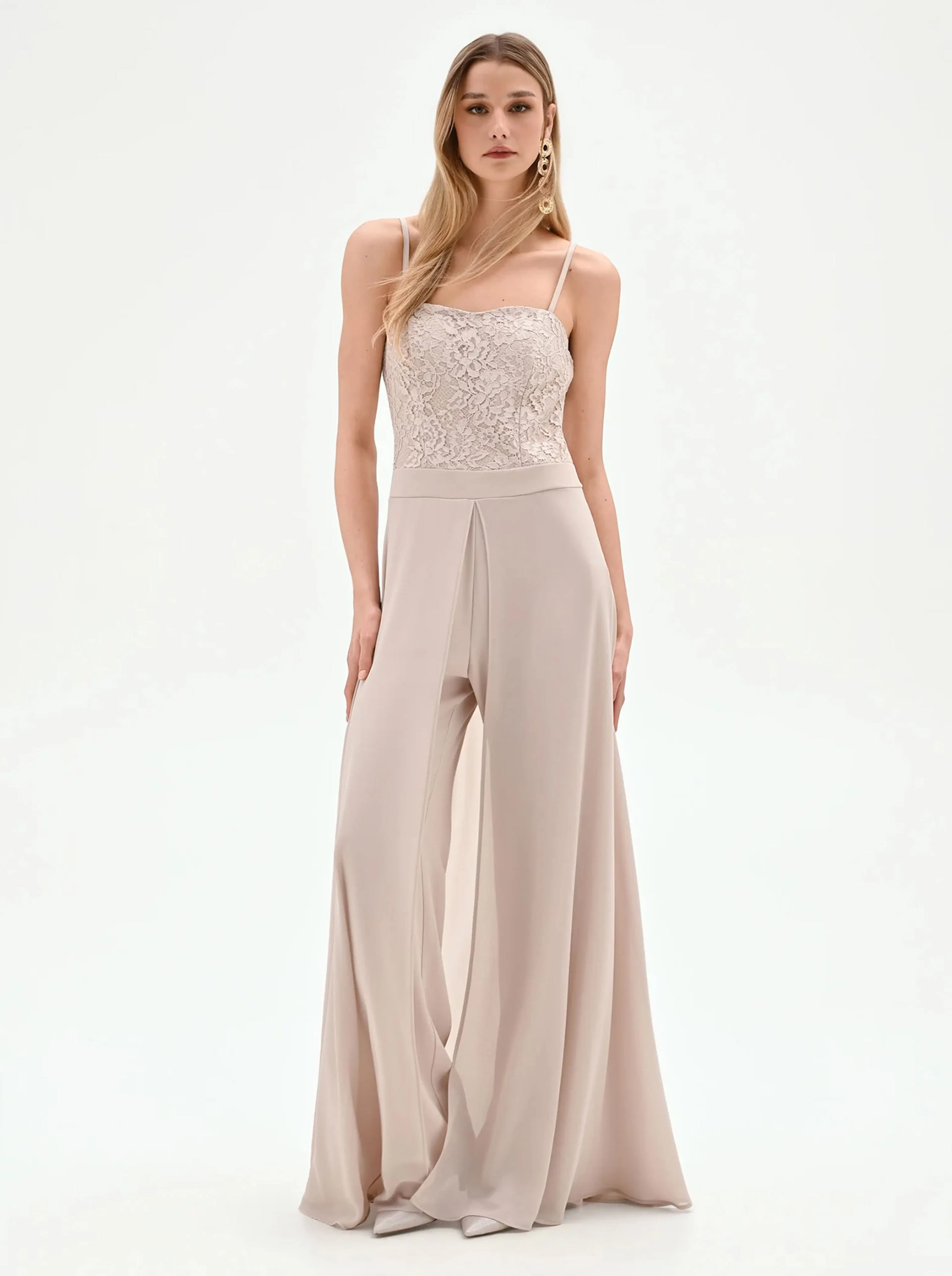 A woman stands confidently in a beige jumpsuit with a delicate lace bodice and flowing wide-leg pants. The neutral tones convey elegance and sophistication.