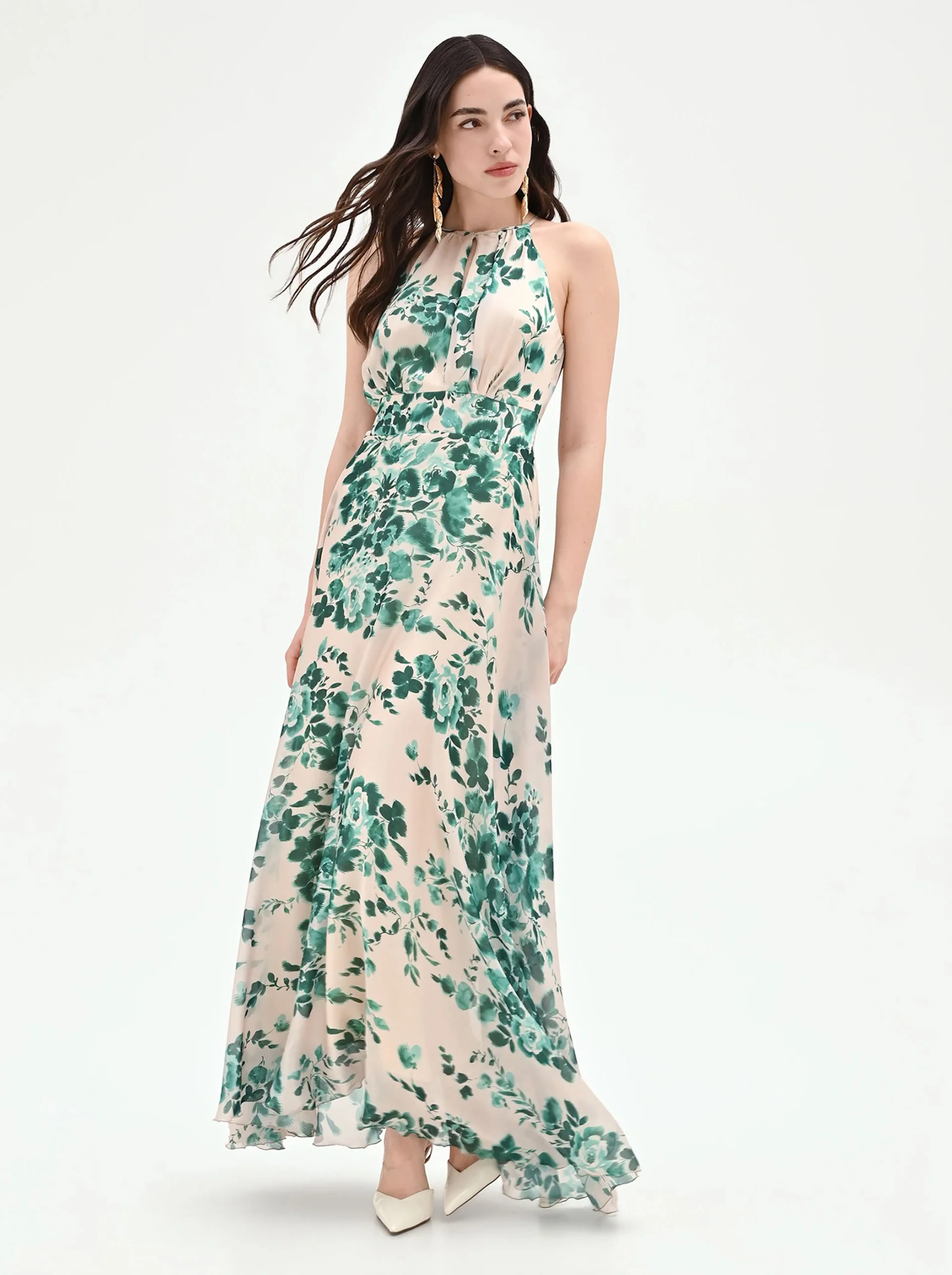 Woman in a flowing halter-neck gown with green floral patterns, standing on a white background. Her hair is loose, conveying elegance and grace.