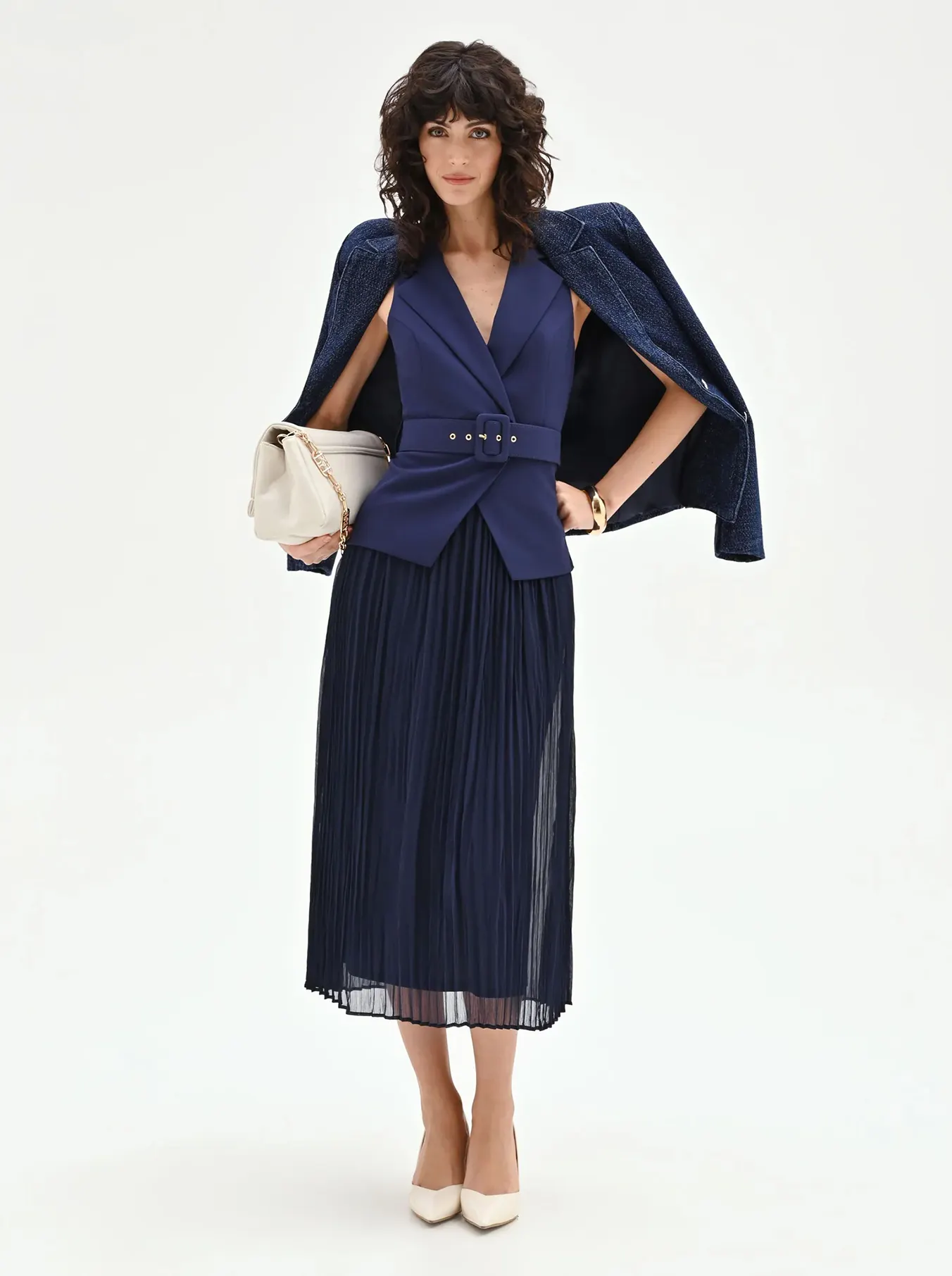 Woman standing confidently in a stylish blue pleated skirt and belted vest, draped with a coat over her shoulders, holding a cream handbag.