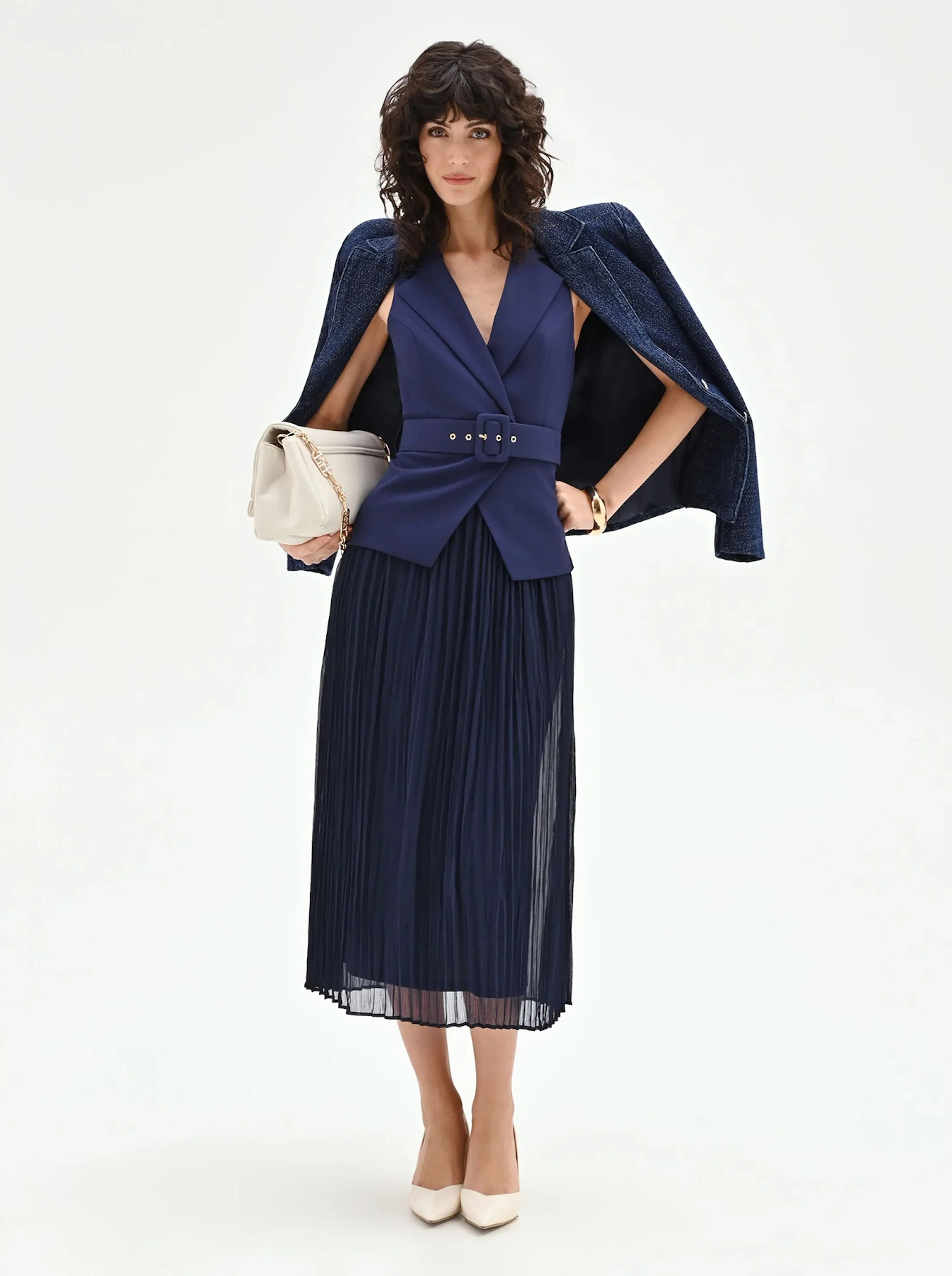 Woman standing confidently in a stylish blue pleated skirt and belted vest, draped with a coat over her shoulders, holding a cream handbag.