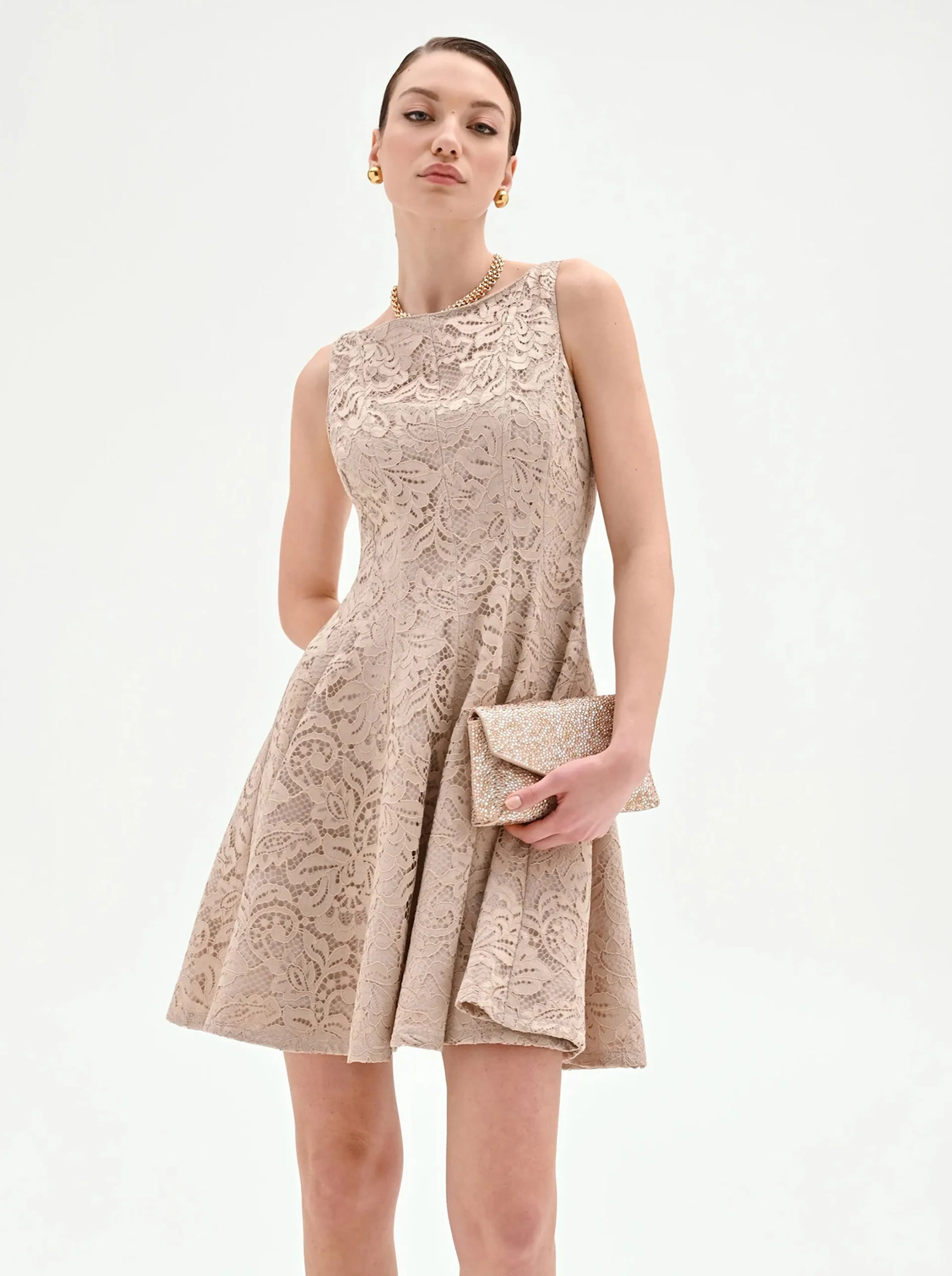 Woman in an elegant beige lace dress holds a sparkling clutch. Her expression is poised, conveying sophistication against a white background.
