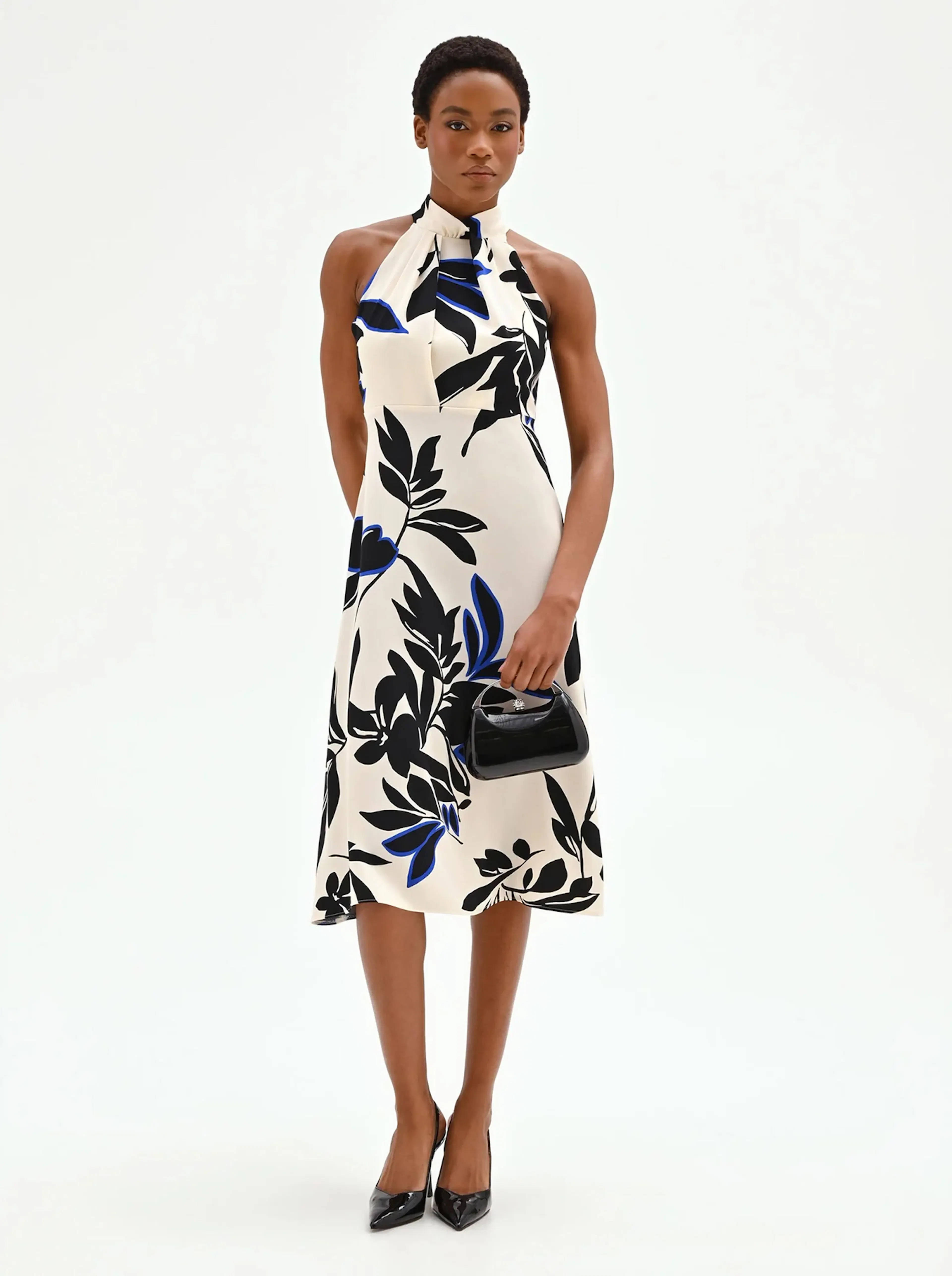 Woman in a halter-neck dress with black and blue floral patterns on white stands confidently against a plain background, holding a small black handbag.
