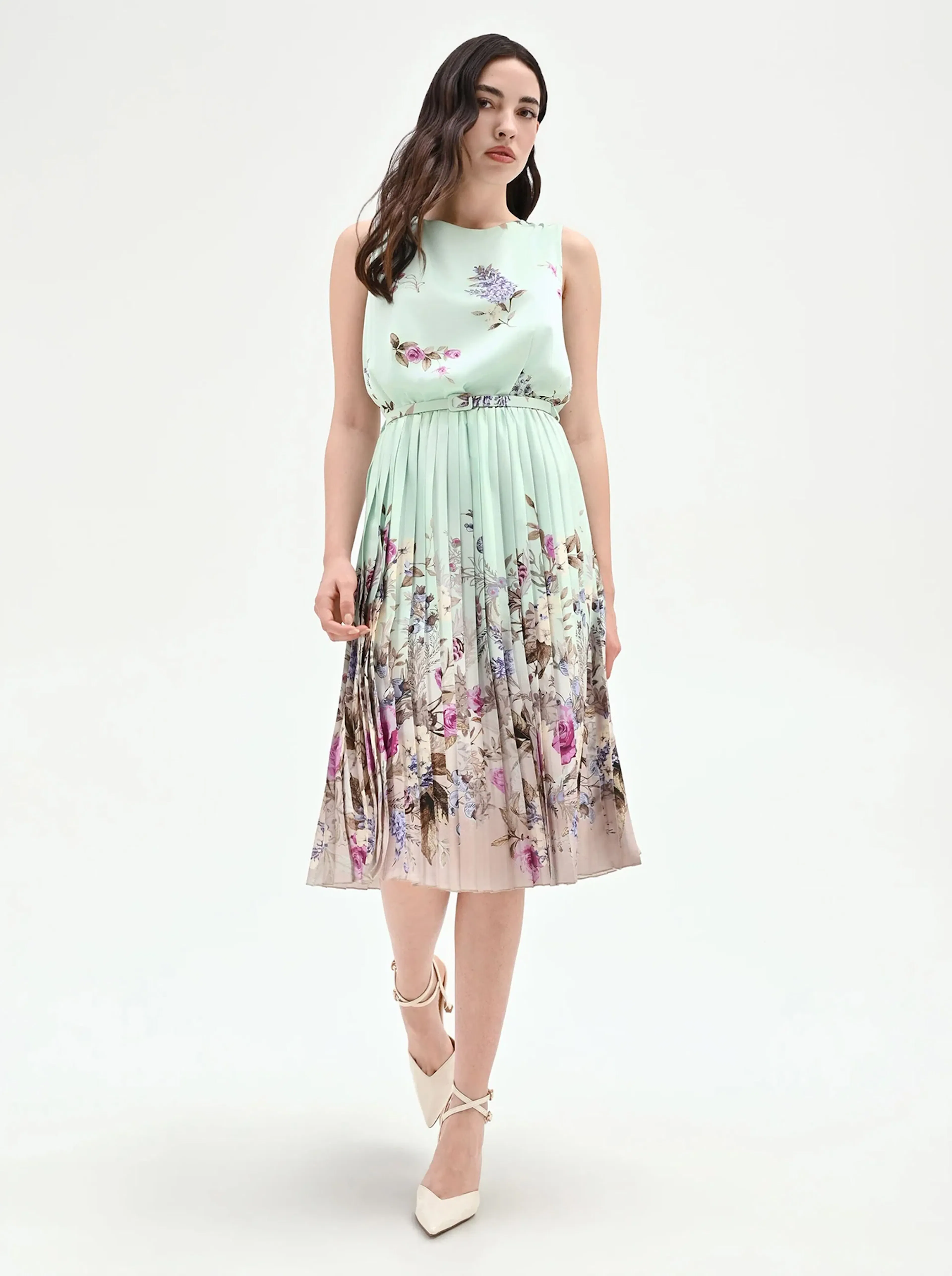 Woman in a sleeveless, mint green dress with floral print stands against white background. She wears white heels, expressing elegance and poise.