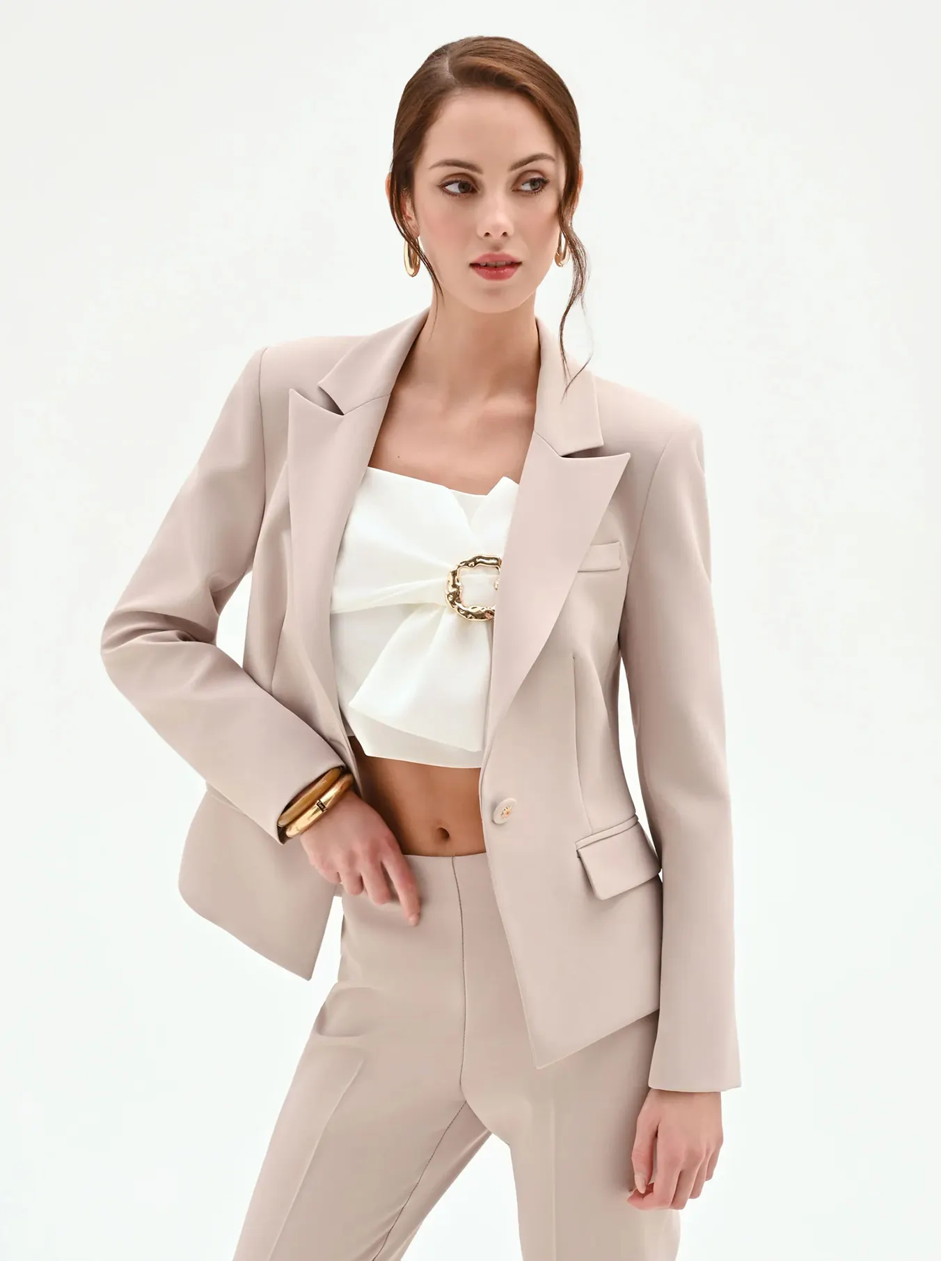 A woman in a beige suit poses confidently against a plain background. She wears a white crop top with a decorative buckle, exuding elegance and sophistication.
