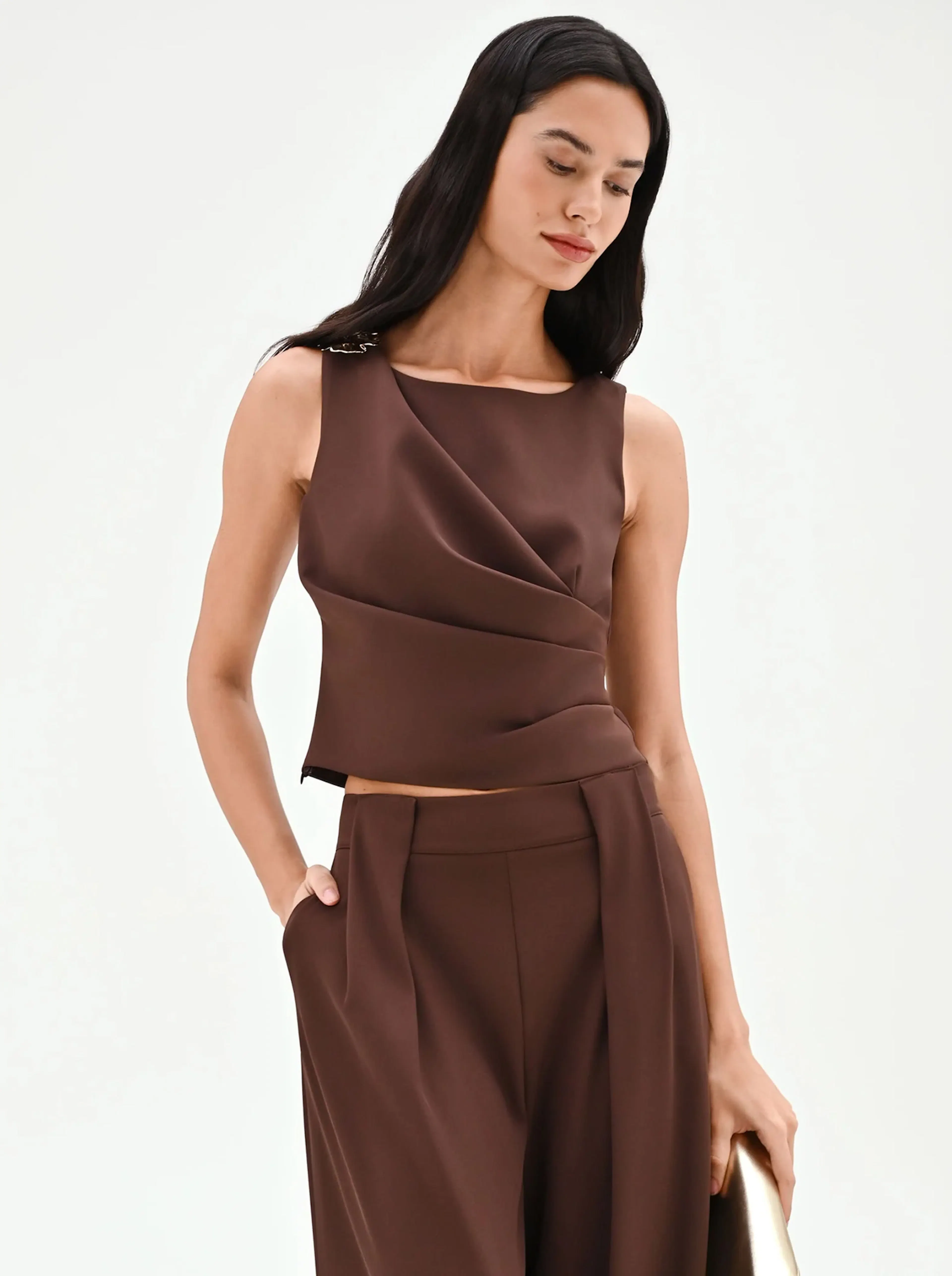 A woman in a chic, sleeveless brown top and matching wide-leg pants stands against a white background, exuding elegance and confidence.
