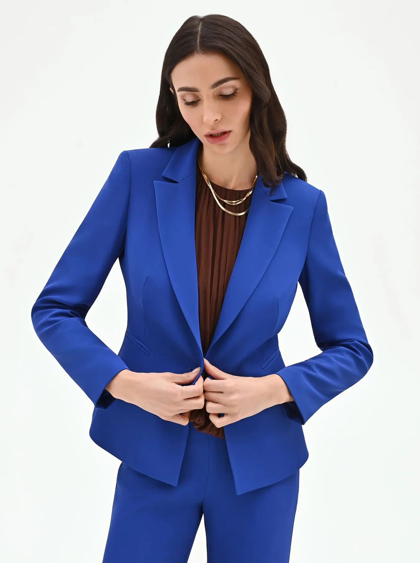 A person with long dark hair wearing a vibrant blue suit over a brown shirt, fastening the jacket with a composed expression against a white background.