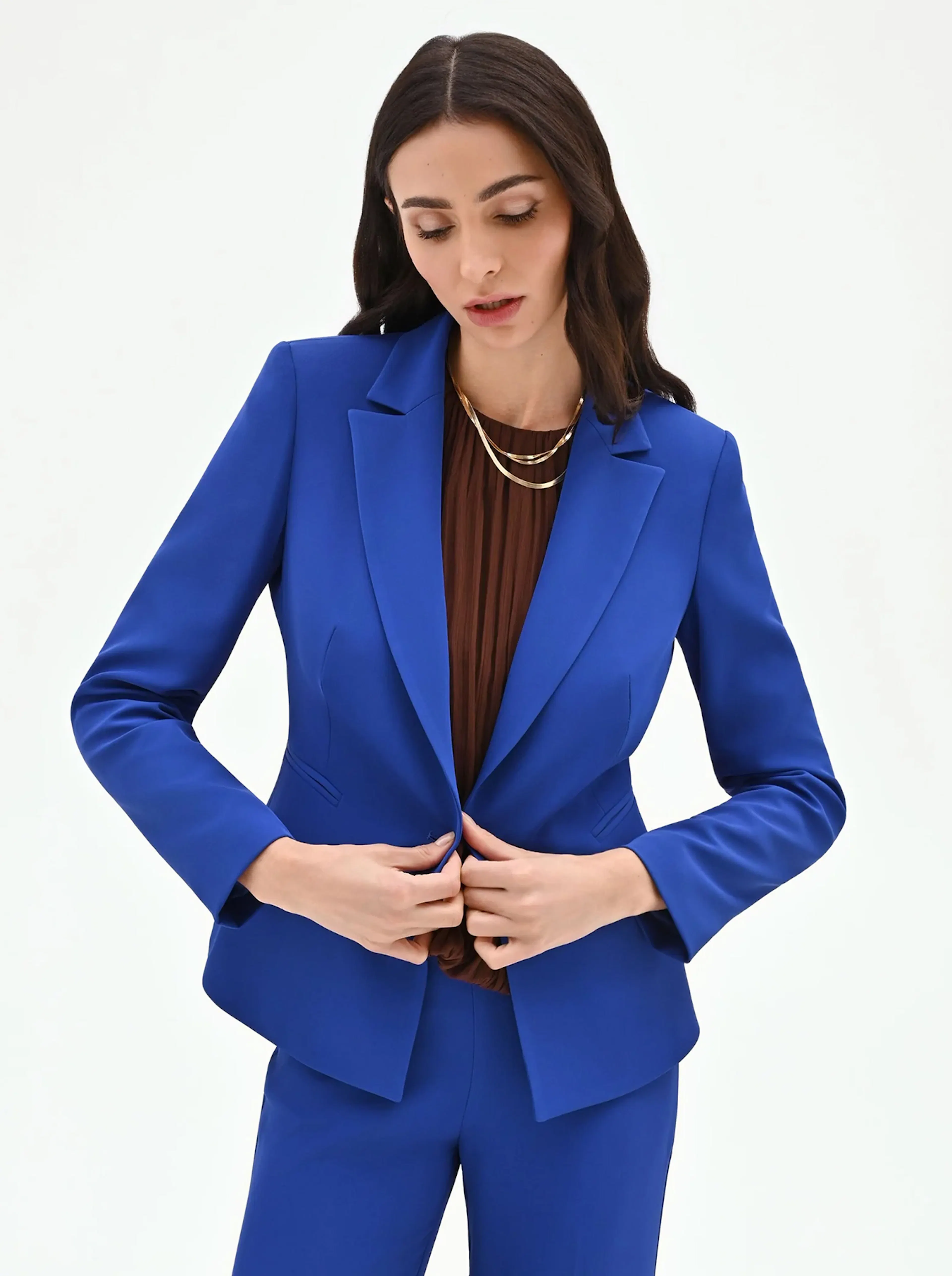 A person with long dark hair wearing a vibrant blue suit over a brown shirt, fastening the jacket with a composed expression against a white background.