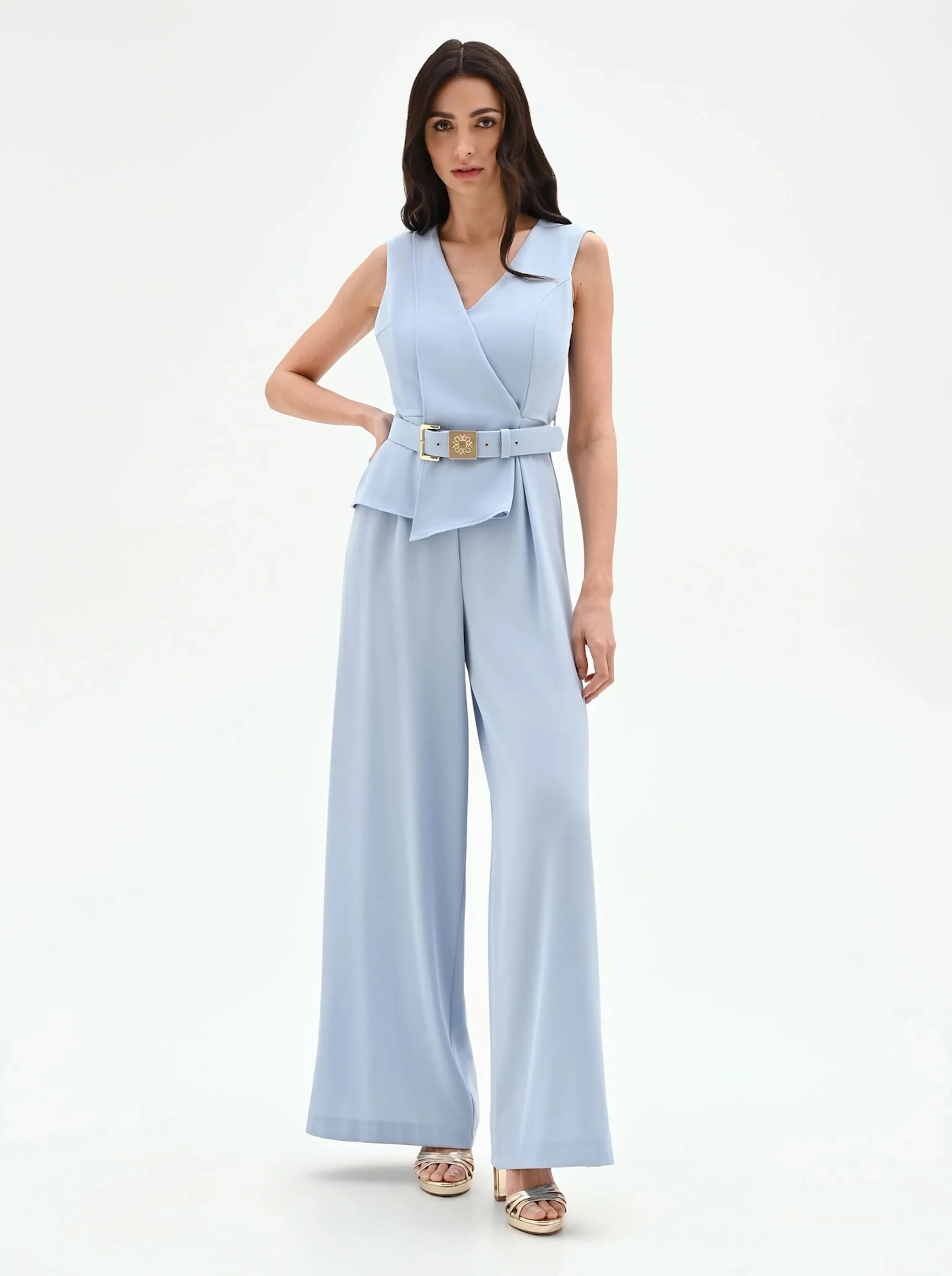A woman poses confidently in a sleeveless, light blue jumpsuit with a wrap-style top and wide-legged pants, accessorized with a belt. She stands against a plain white background, exuding elegance and poise.