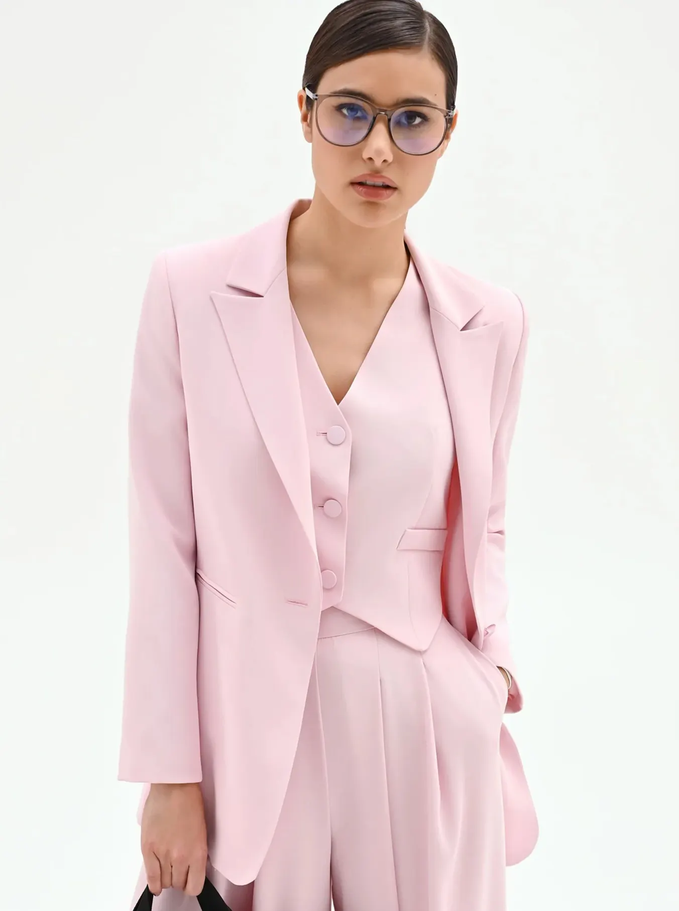 Young woman in a pale pink suit with a blazer, vest, and wide-leg pants stands confidently. She wears stylish glasses and carries a black handbag.