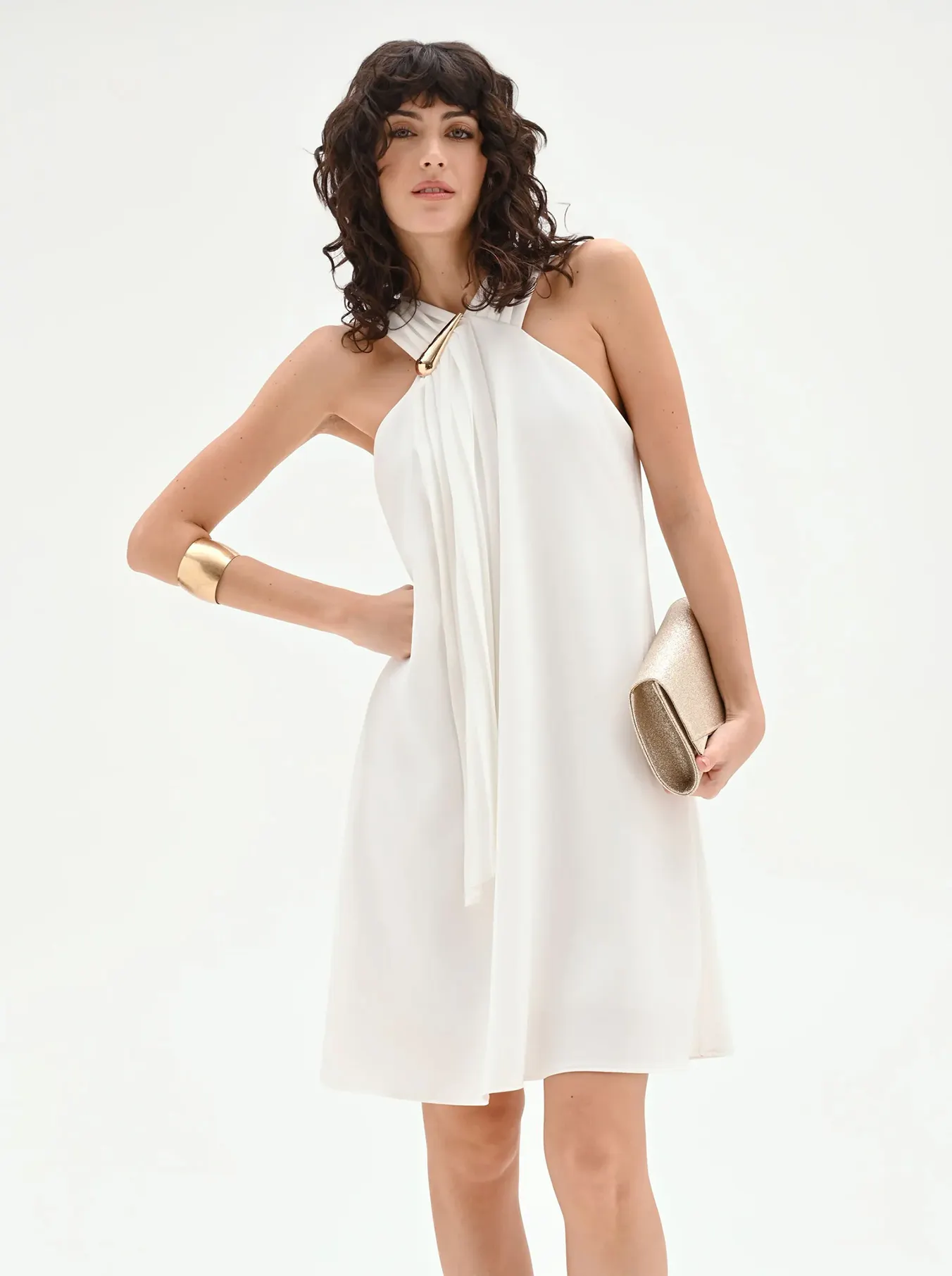Woman in a stylish sleeveless white halter dress with gold accessories and clutch, exuding elegance and confidence against a white background.