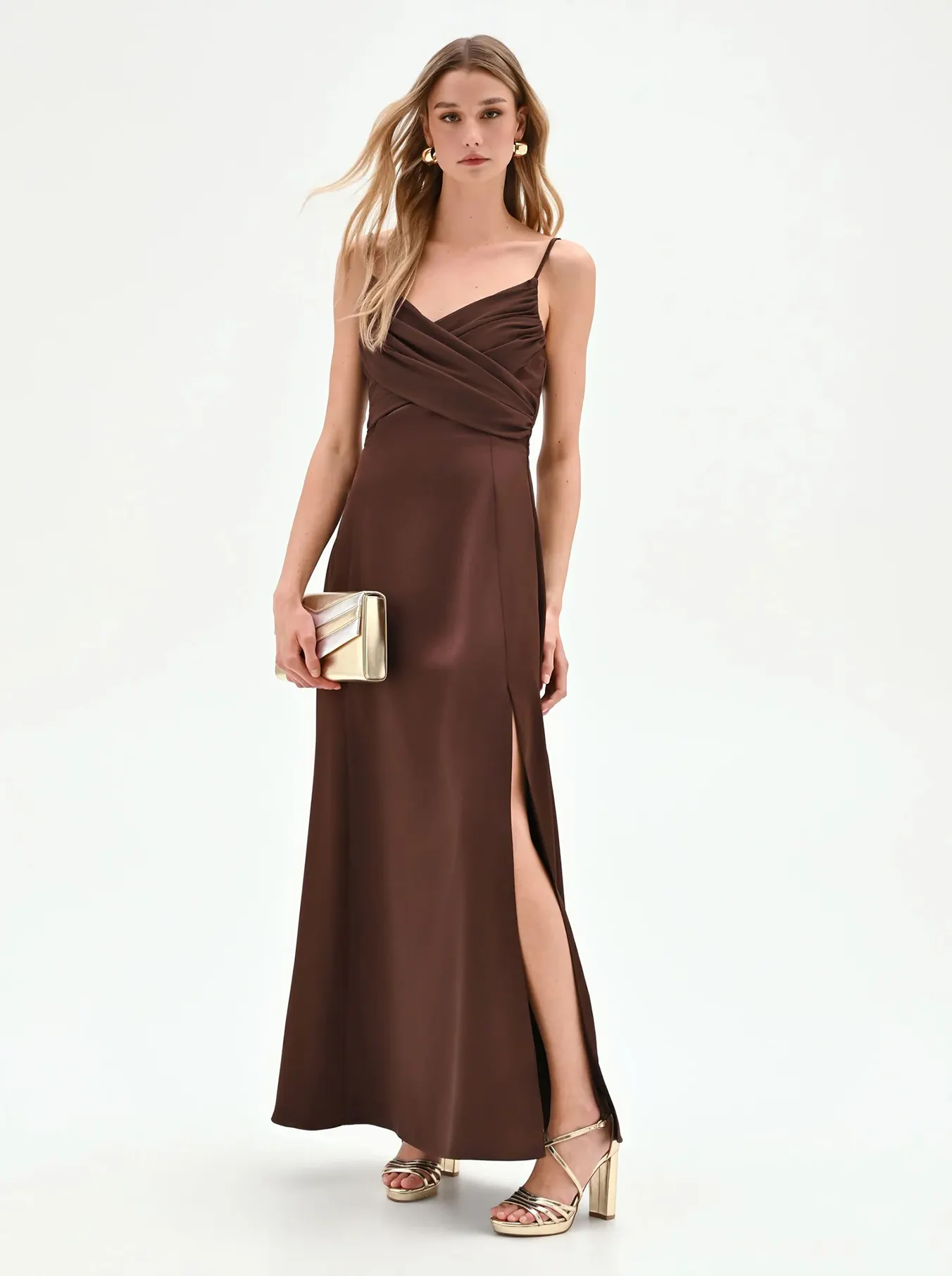 A woman in a chocolate brown, sleeveless satin gown with a thigh-high slit stands gracefully. She holds a gold clutch and wears matching heels, exuding elegance.