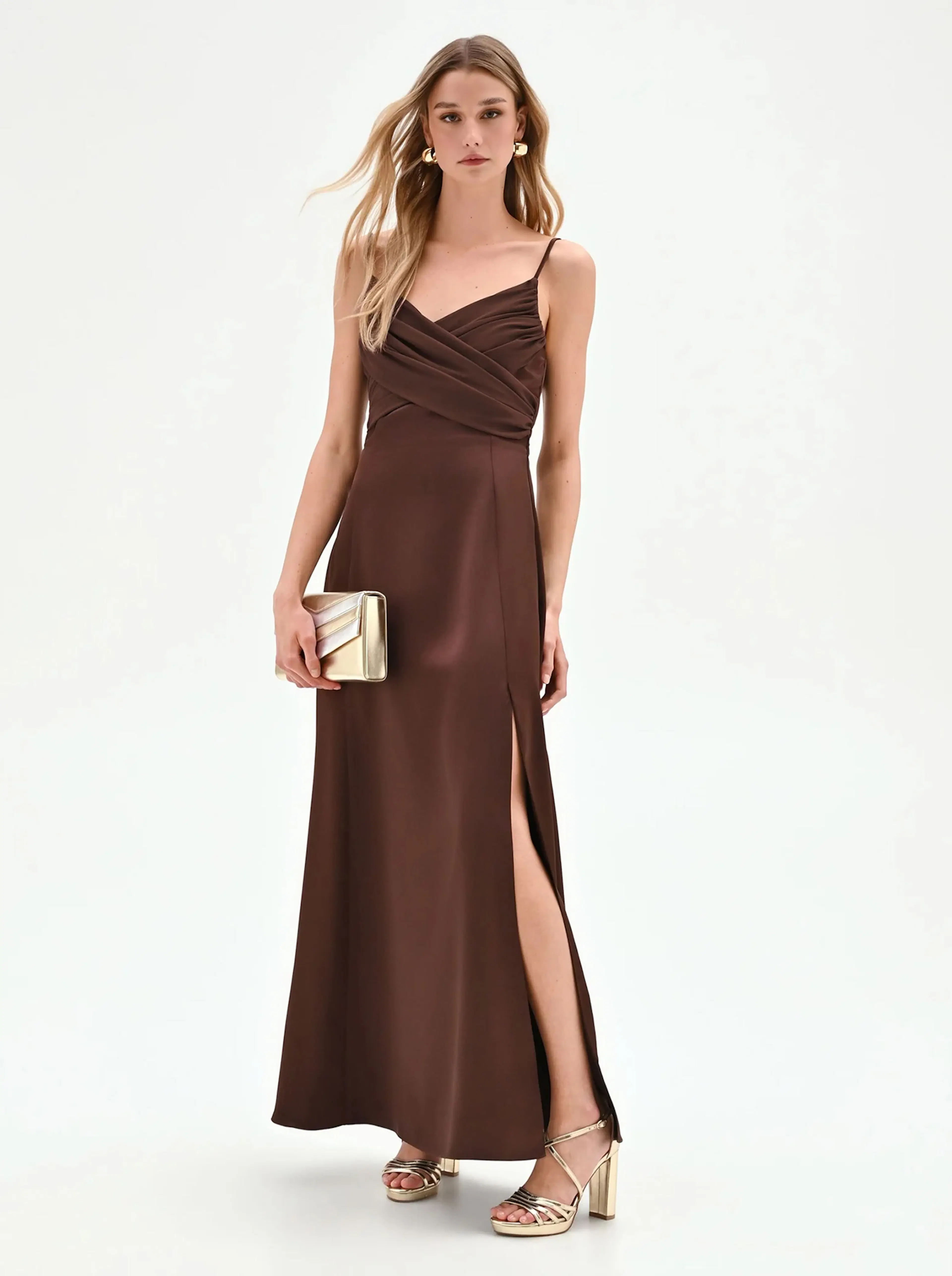 A woman in a chocolate brown, sleeveless satin gown with a thigh-high slit stands gracefully. She holds a gold clutch and wears matching heels, exuding elegance.