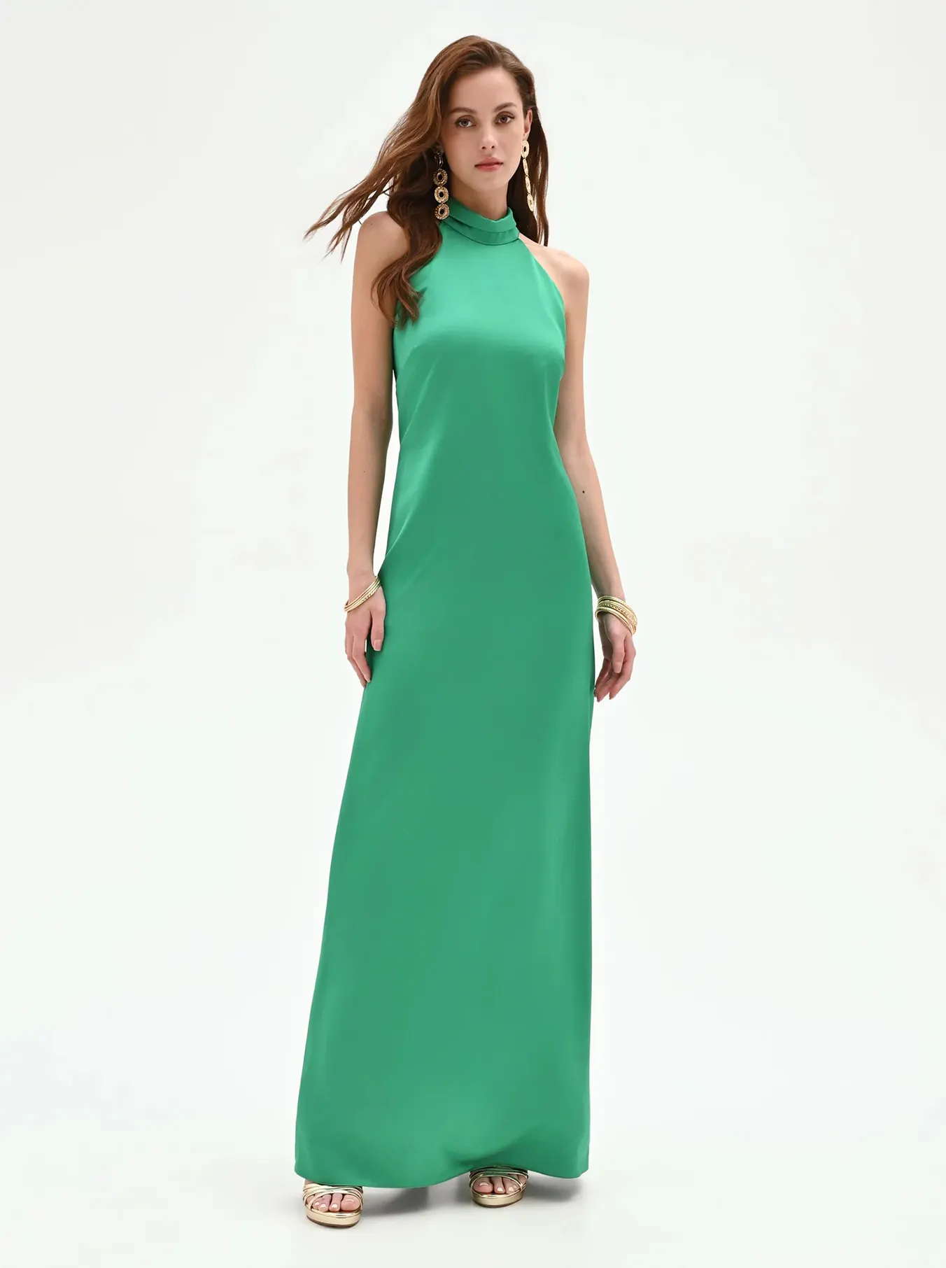 Woman in a green halter-neck gown, standing against a white background. She wears gold earrings and bangles, exuding elegance and confidence.