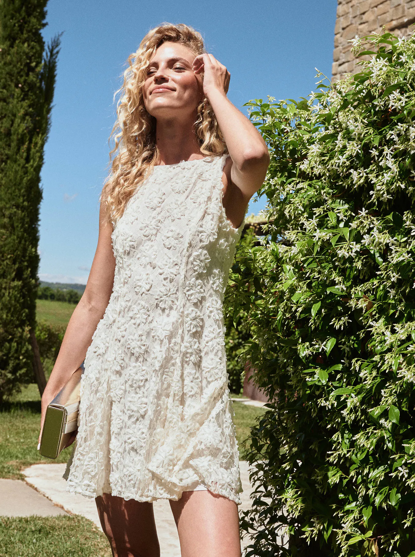 A woman with curly blonde hair wearing a white lace dress smiles in a sunny garden. She holds a book, with lush greenery and a stone building nearby.