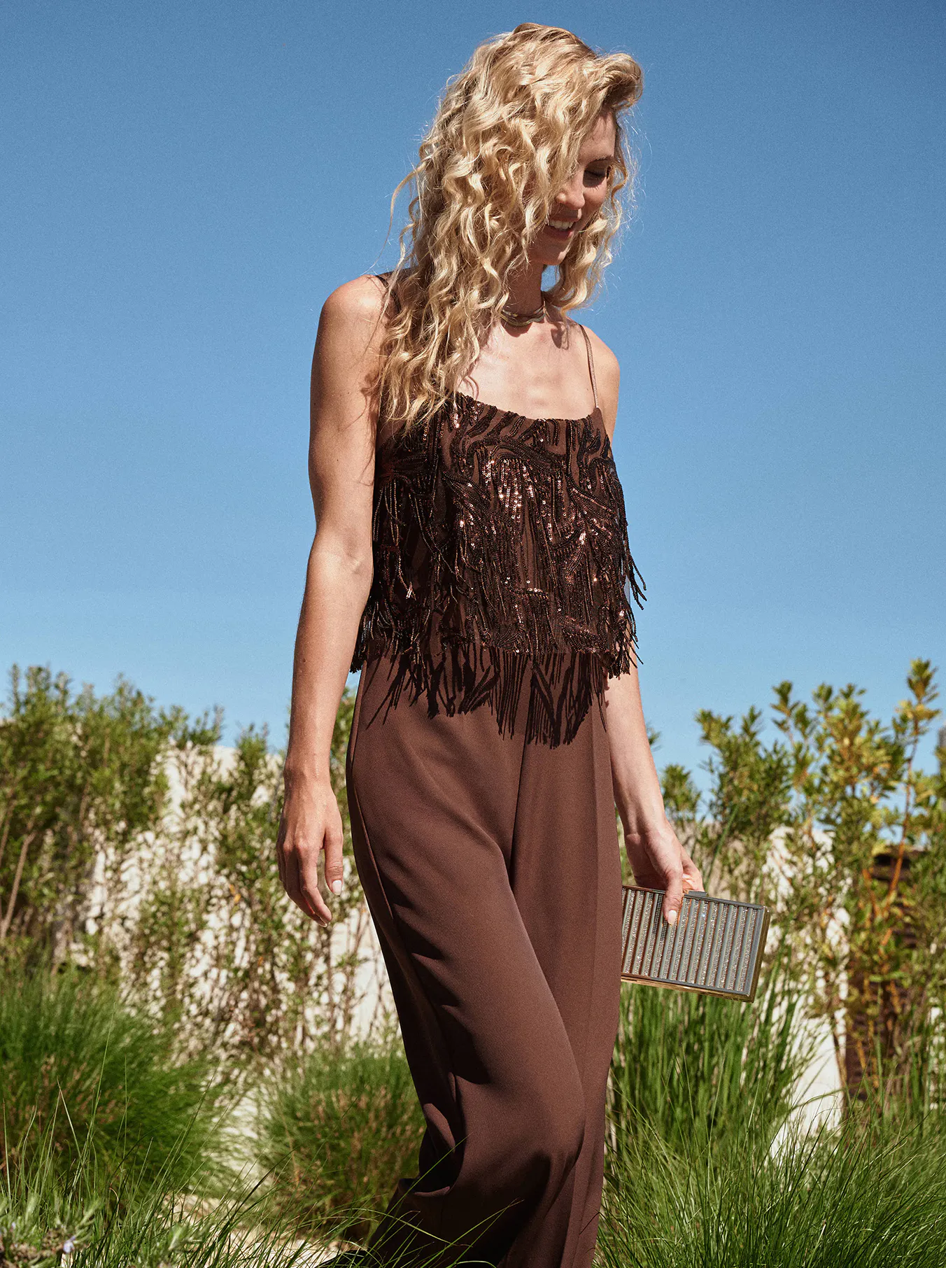 A woman with curly blonde hair wears a brown fringe top and pants, holding a metallic clutch. She walks in a sunny garden, exuding elegance.