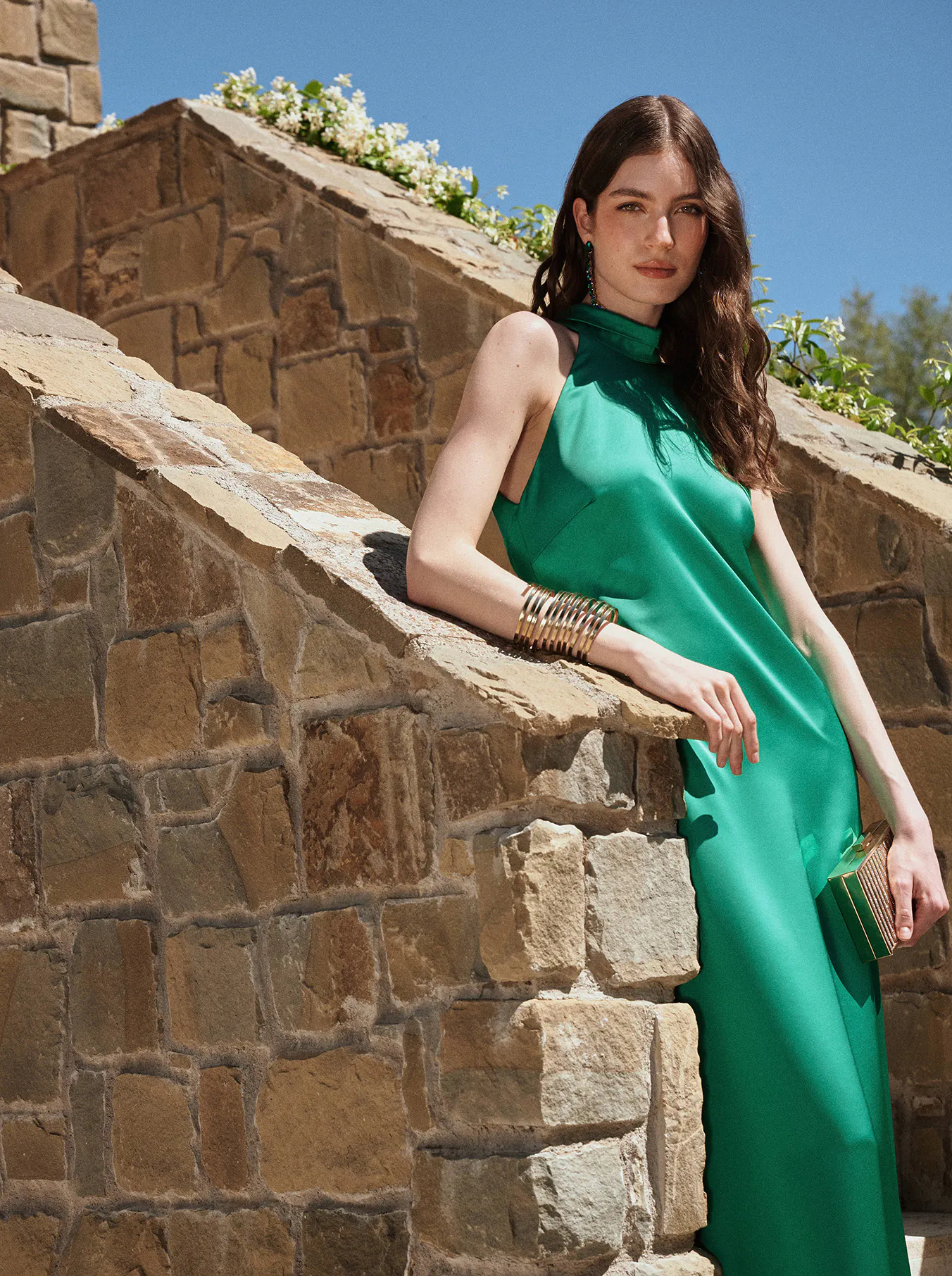 A woman in a flowing green dress leans against a stone wall under a clear blue sky. She wears gold bracelets, exuding elegance and tranquility.