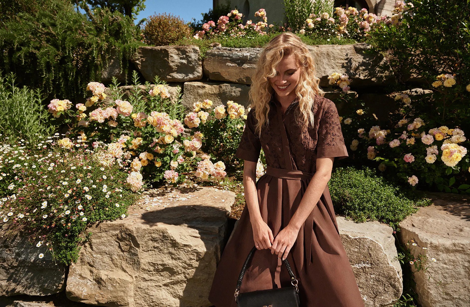 Woman in a brown dress sitting on a rock surrounded by blooming pink and yellow roses in a sunny garden.