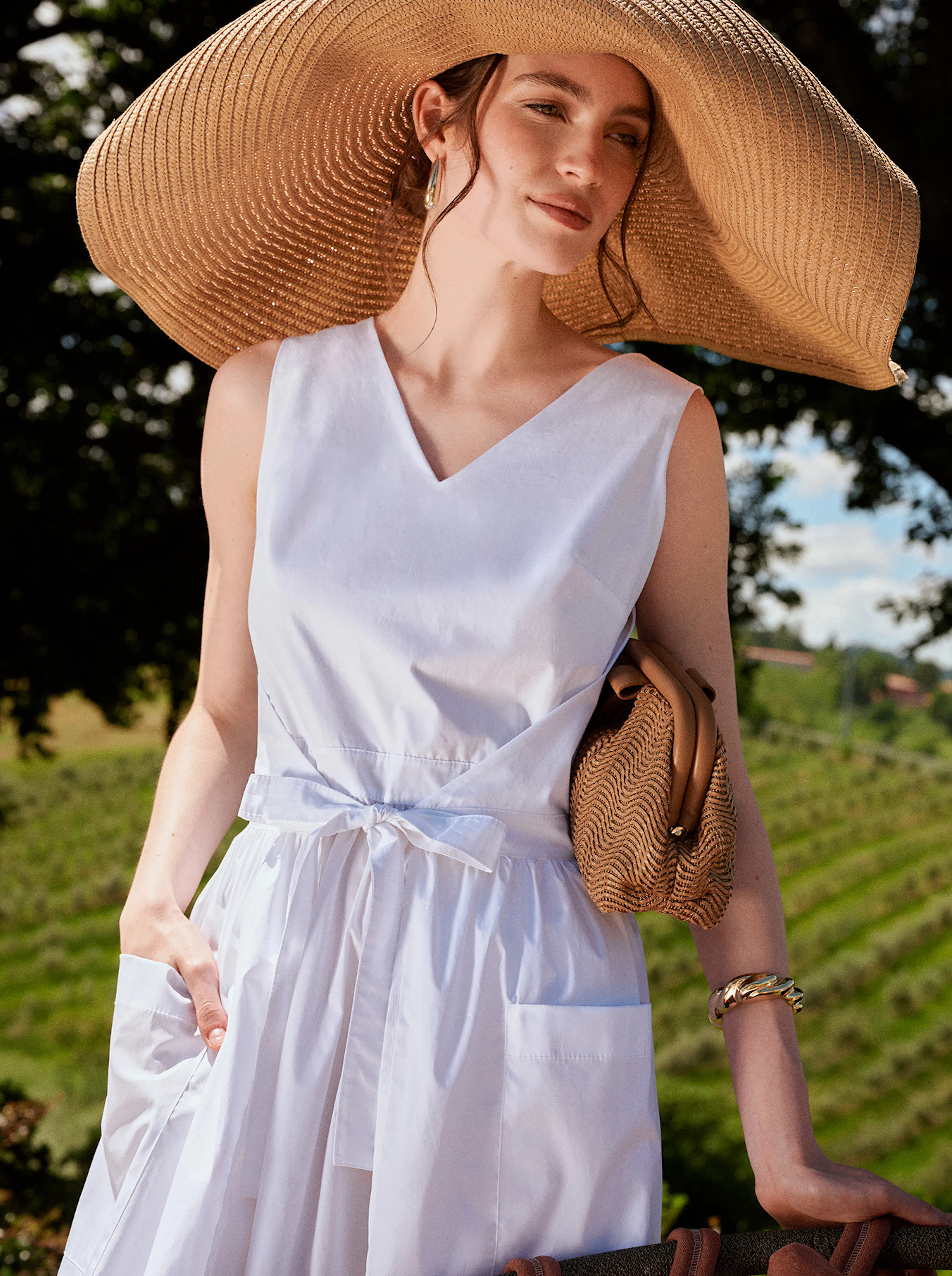 Woman in a white sleeveless dress and large straw hat holding a brown woven handbag outdoors in a sunny, green vineyard setting.