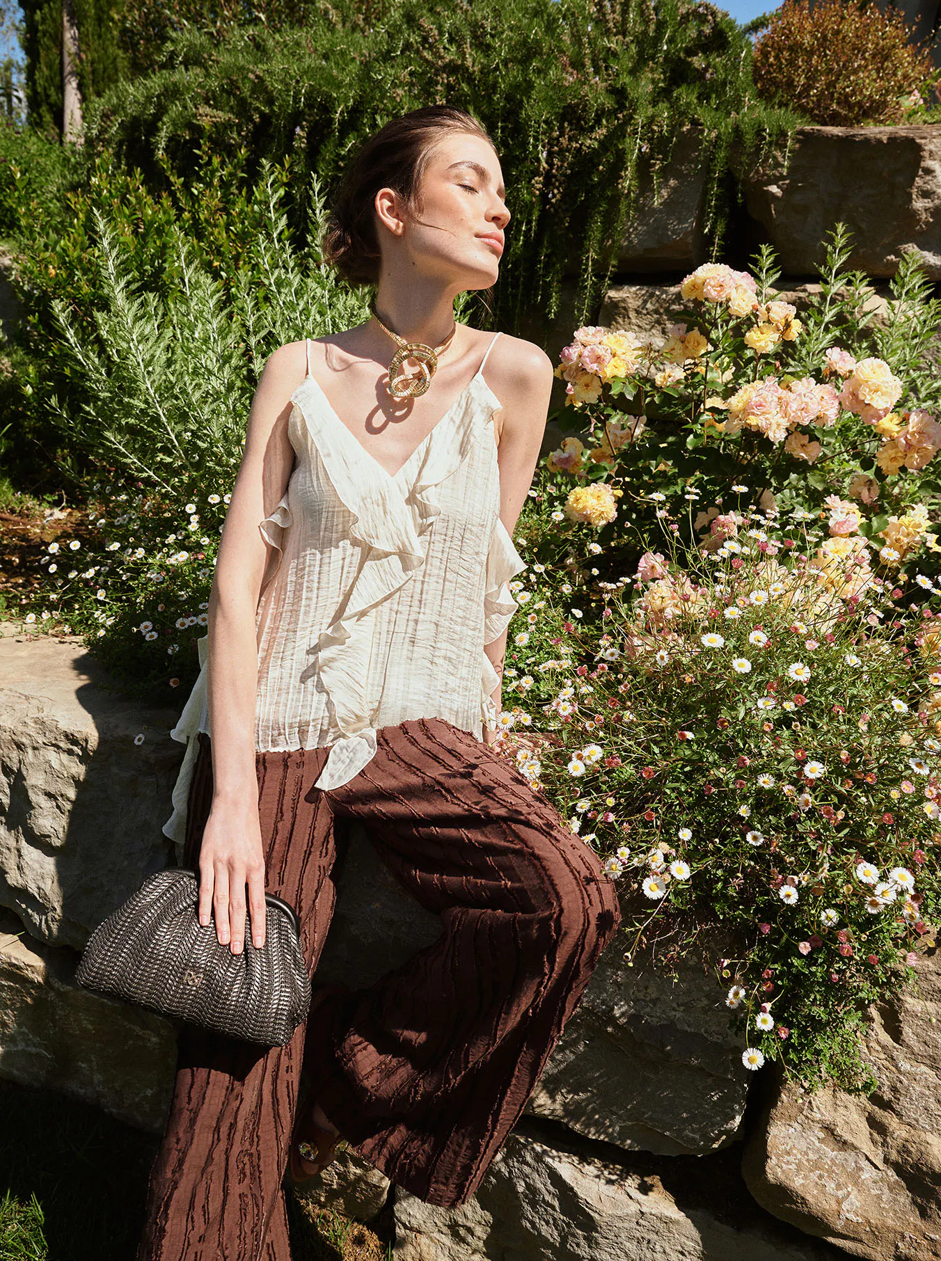 A woman in a white ruffled top and brown pants sits on a stone wall, basking in the sun. She's holding a textured clutch, surrounded by lush greenery and blooming flowers.