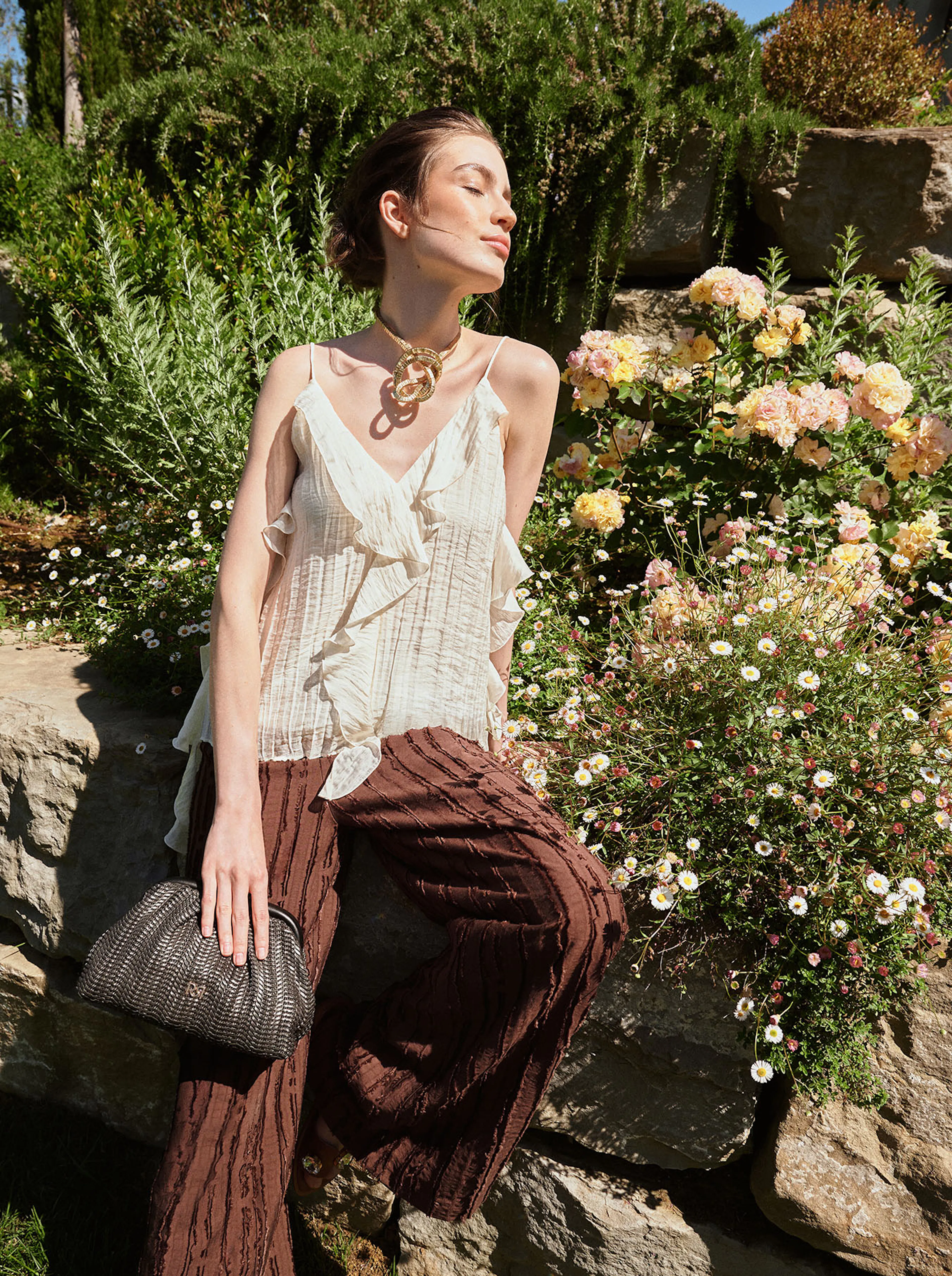 A woman in a white ruffled top and brown pants sits on a stone wall, basking in the sun. She's holding a textured clutch, surrounded by lush greenery and blooming flowers.