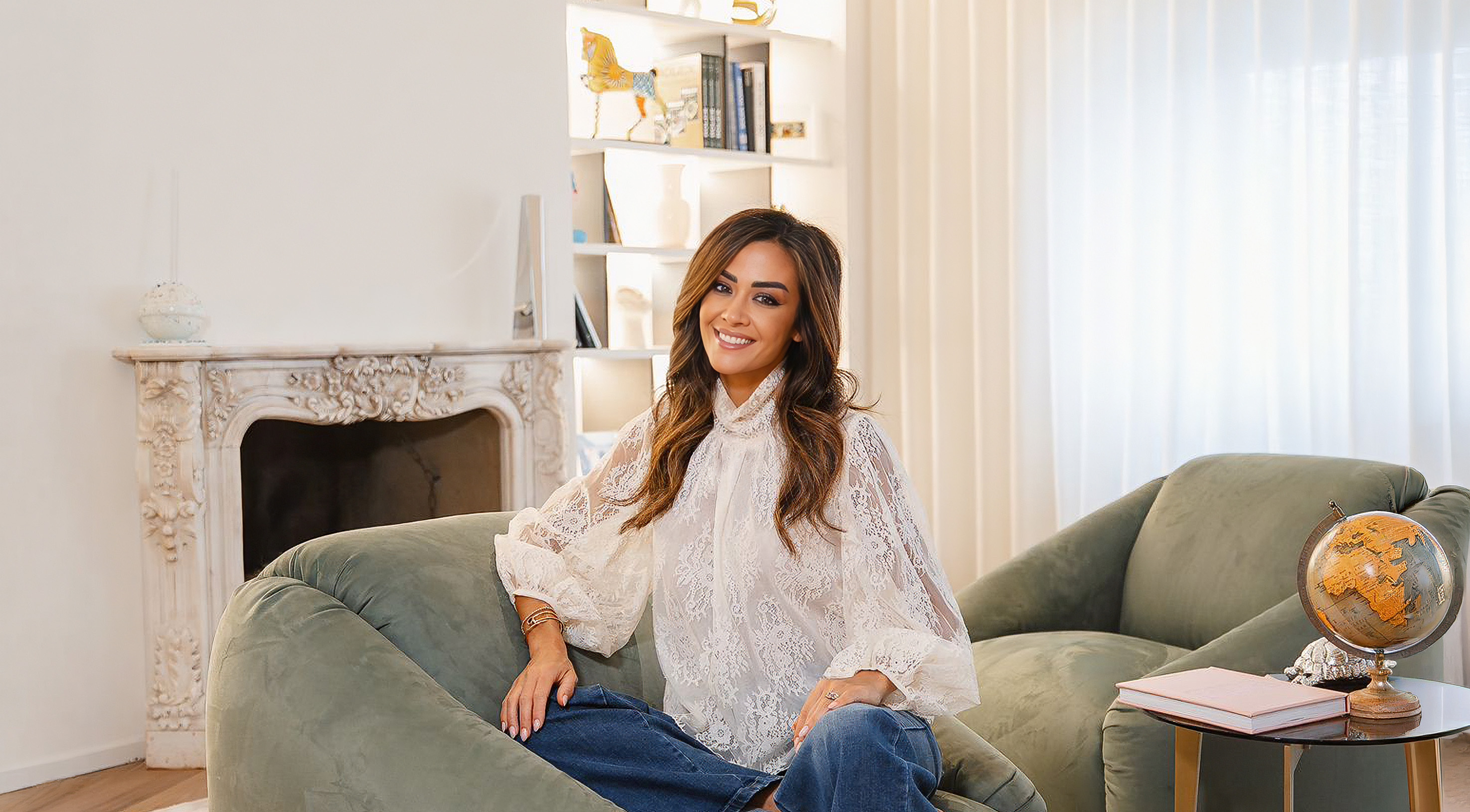 Giorgia Palmas in white lace blouse and blue jeans sitting on a green sofa in a bright living room with a globe and fireplace.