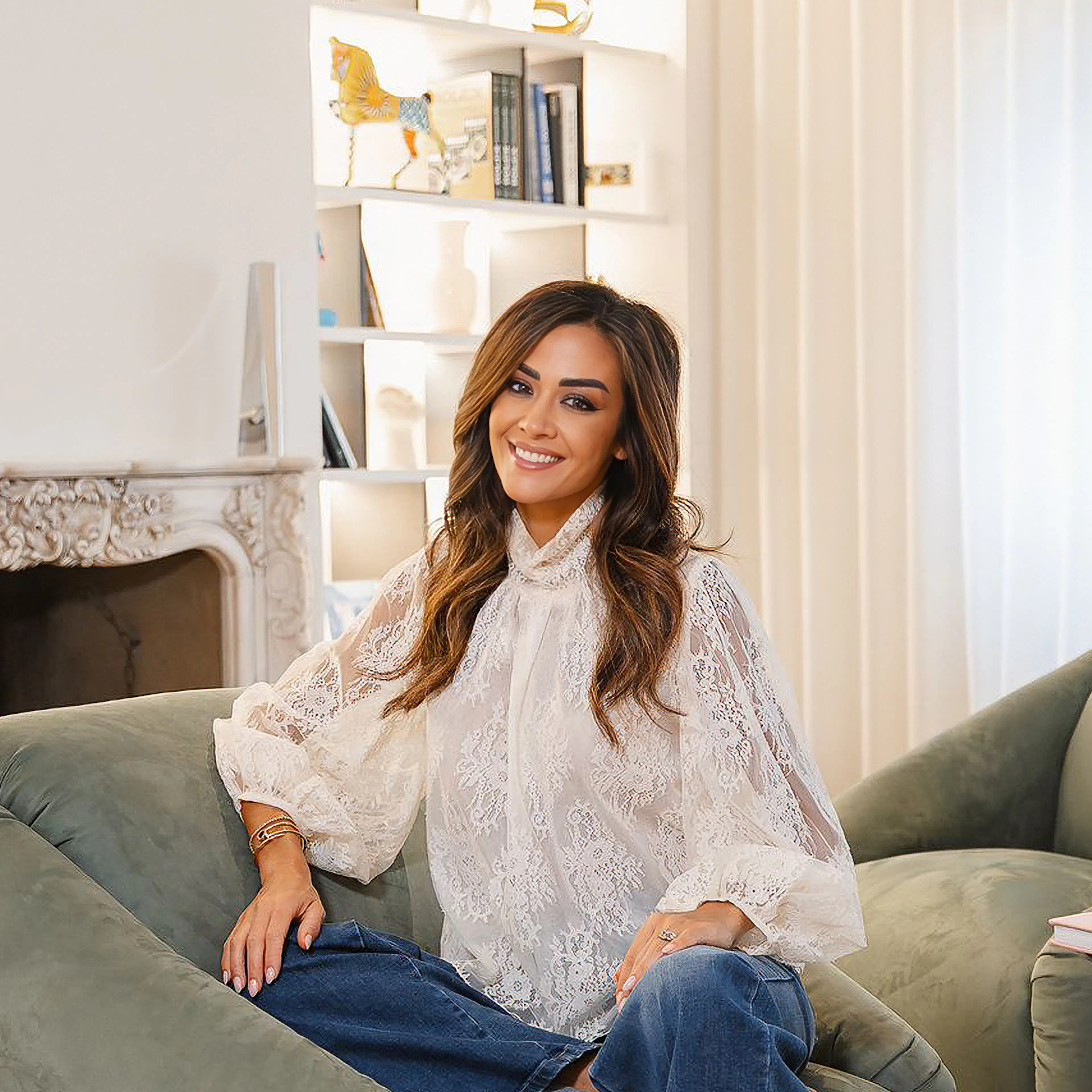 Giorgia Palmas in white lace blouse and blue jeans sitting on a green sofa in a bright living room with a globe and fireplace.