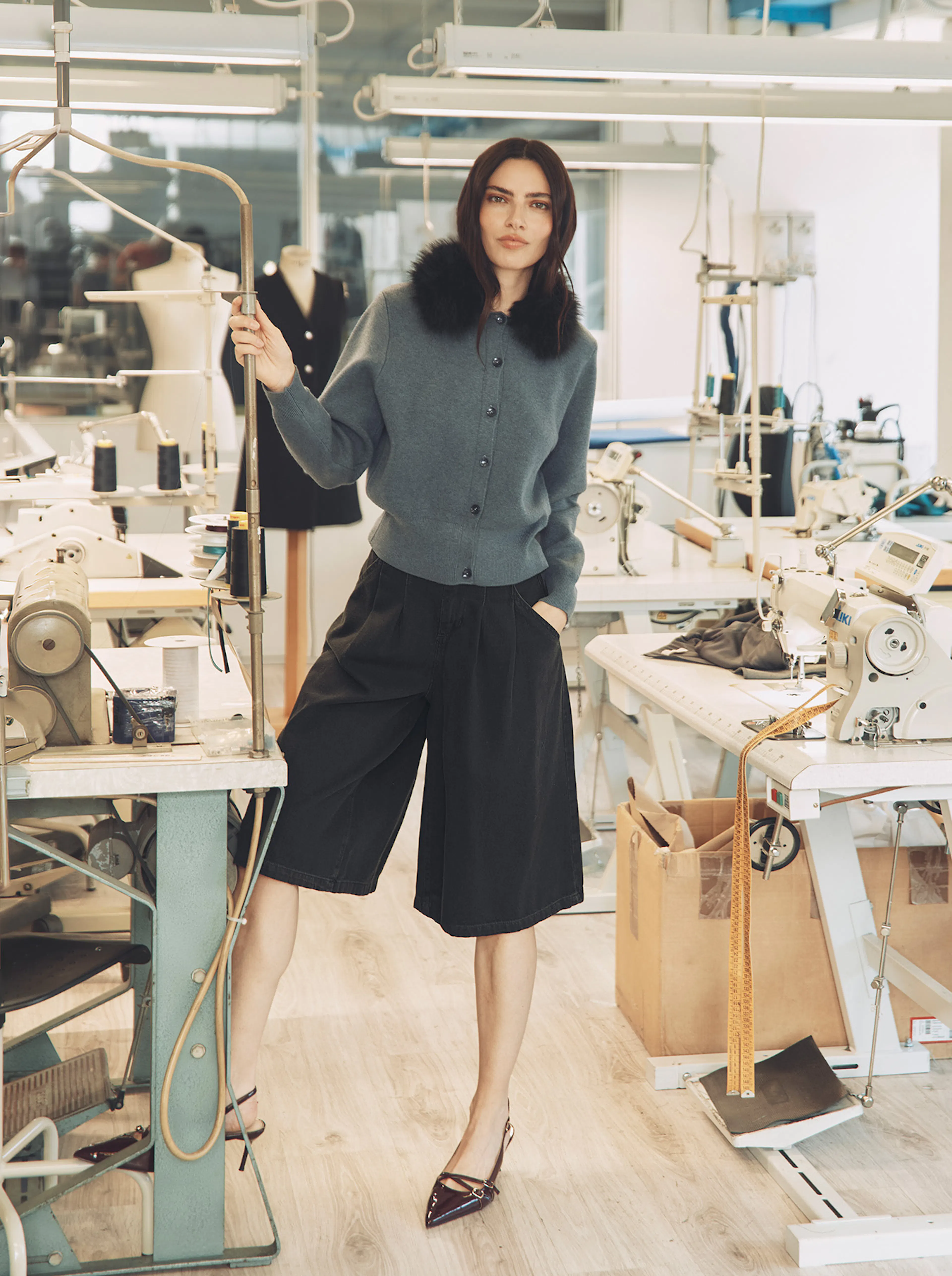 A brunette model poses in a fashion workshop, leaning casually against a metal frame. She is wearing a grey button-down cardigan with a dark faux-fur collar and wide-leg black culottes, finished with burgundy pointed-toe heels. Several sewing machines and a mannequin can be seen behind her.