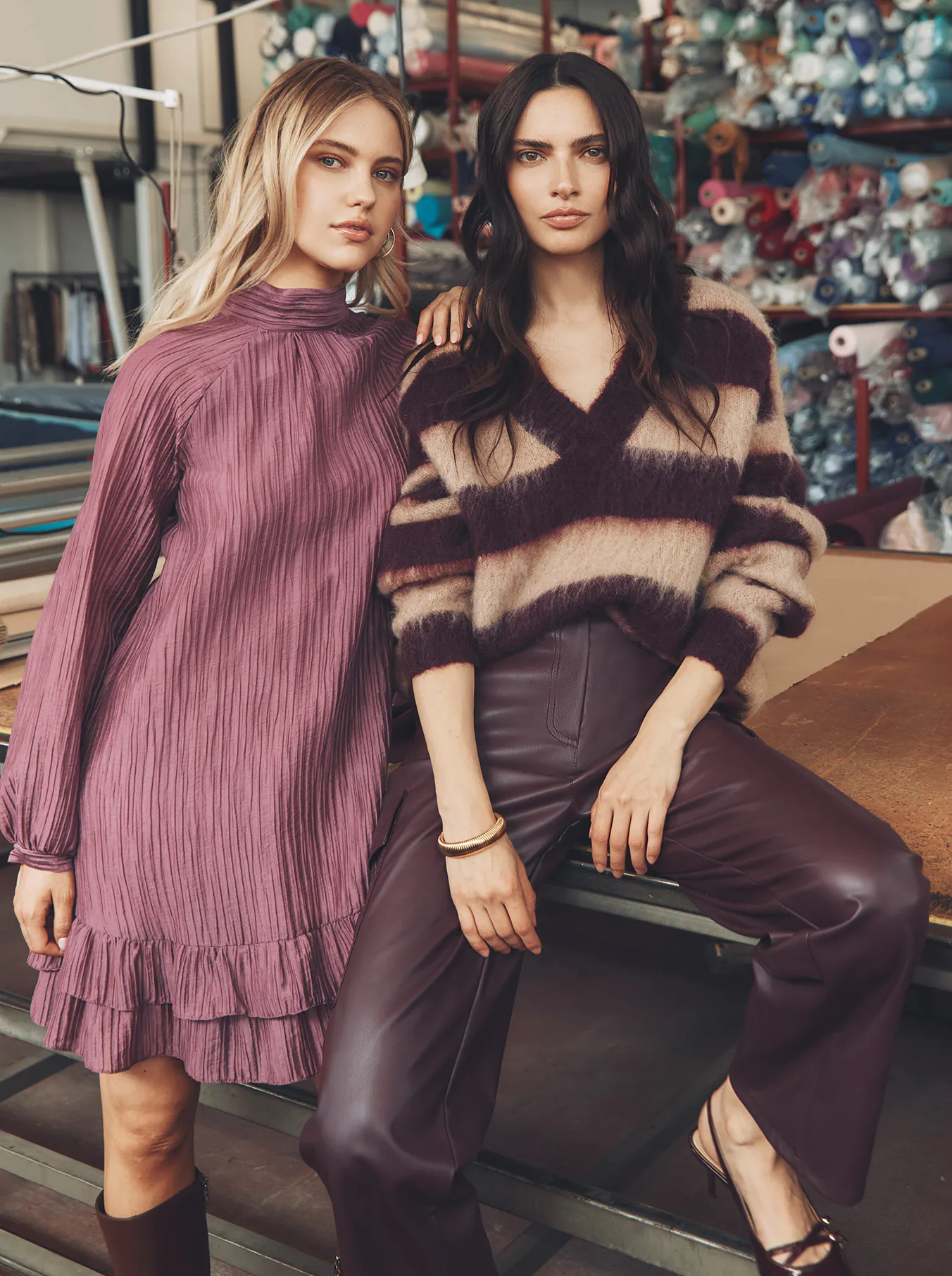 A portrait of two models in a textile factory setting. On the left, a blonde model wears a purple, pleated high-neck midi dress. On the right, a brunette model wears a v-neck, striped mohair sweater with wide-leg burgundy leather trousers. They both look directly at the camera, leaning against a work table. The background is filled with shelves of fabric rolls.