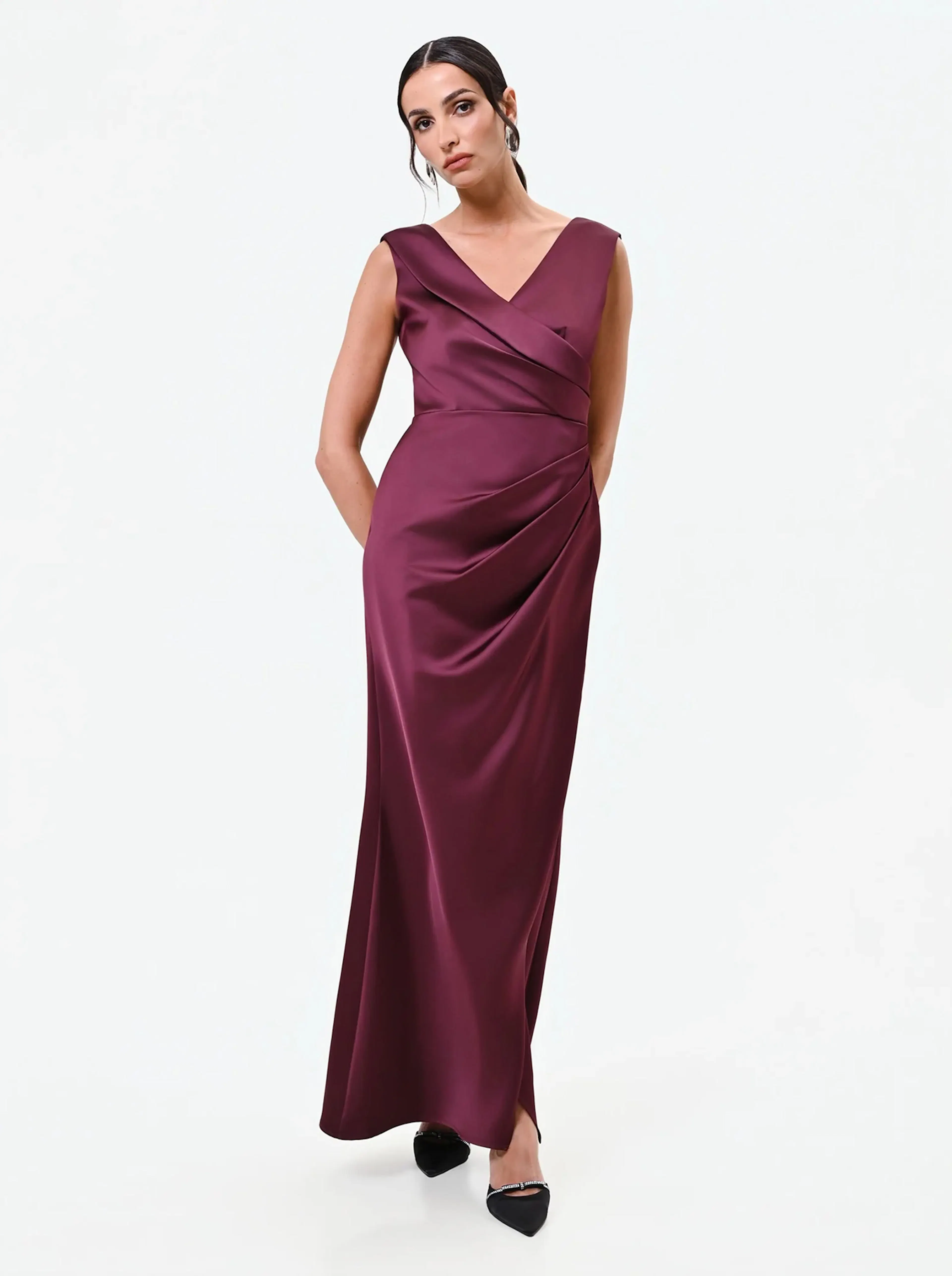 Woman in an elegant, sleeveless burgundy satin gown with a V-neckline and draped detail. She stands confidently against a plain white background.