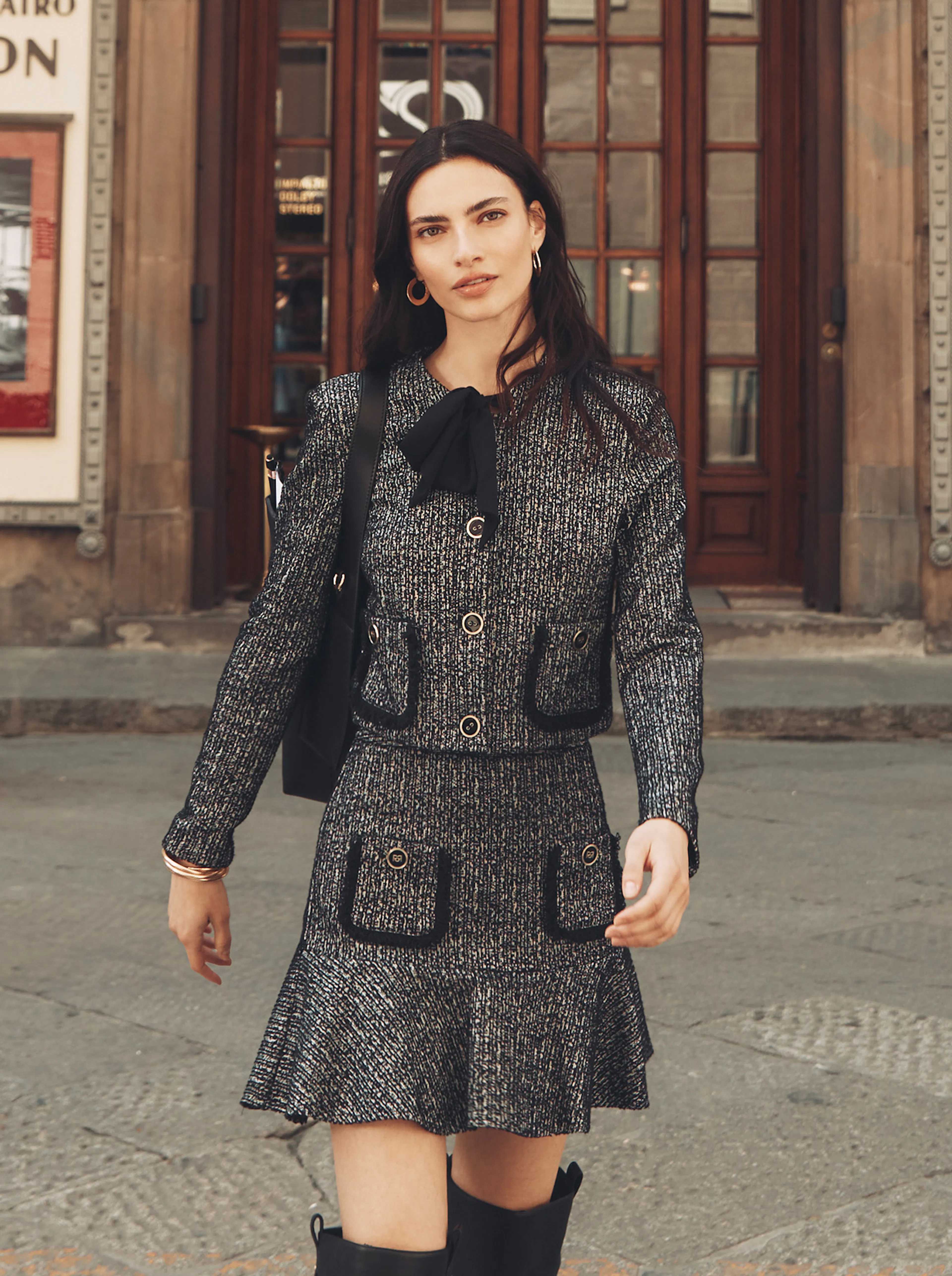 A woman with long dark wavy hair walks down a street in an Italian city, looking directly at the camera. She is wearing a grey, textured tweed skirt suit with a short ruffled skirt and jacket trimmed with dark blue piping and gold buttons, accented by a dark neck bow. She wears large gold hoop earrings and carries a dark bag slung over her shoulder. Historic wooden doors are visible behind her.