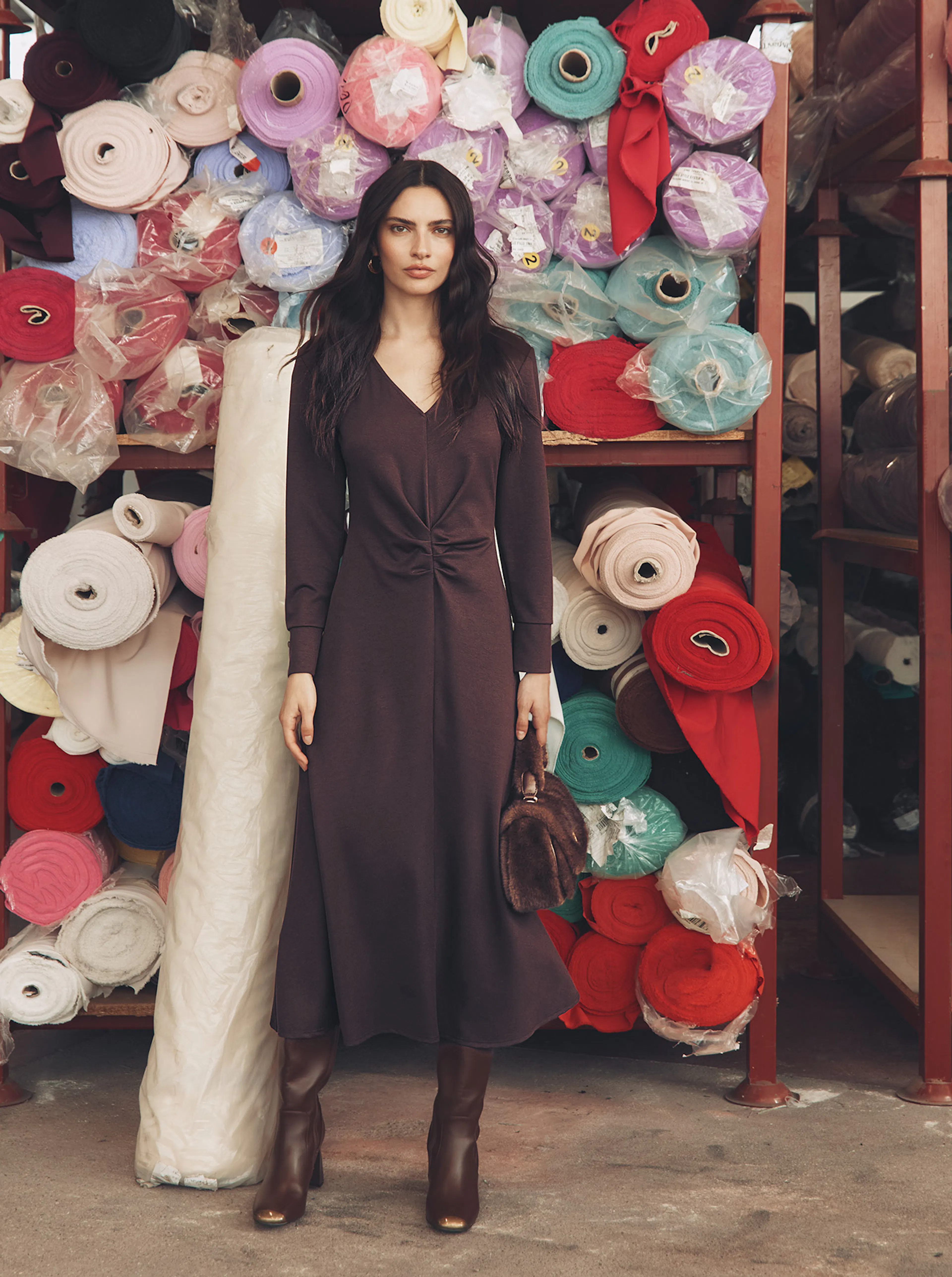 A brunette model stands in a warehouse aisle filled with large shelves stacked with colourful fabric rolls. She is wearing a long-sleeved burgundy midi dress with ruched detailing at the waist, brown knee-high boots, and holds a small brown faux-fur handbag.