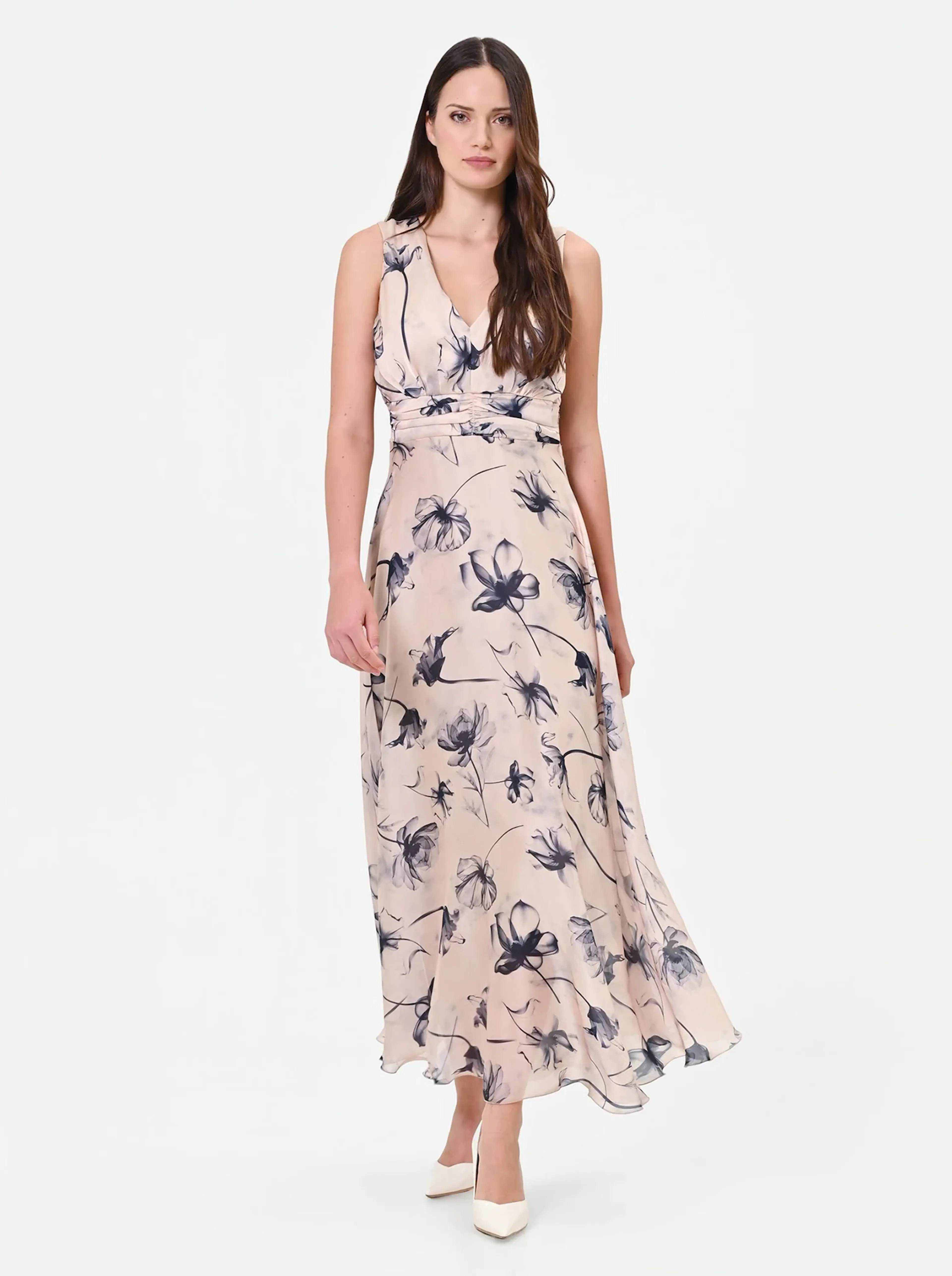 A woman in a sleeveless, floral print maxi dress with a V-neck stands confidently. The dress is light pink with dark floral patterns. She wears white heels.