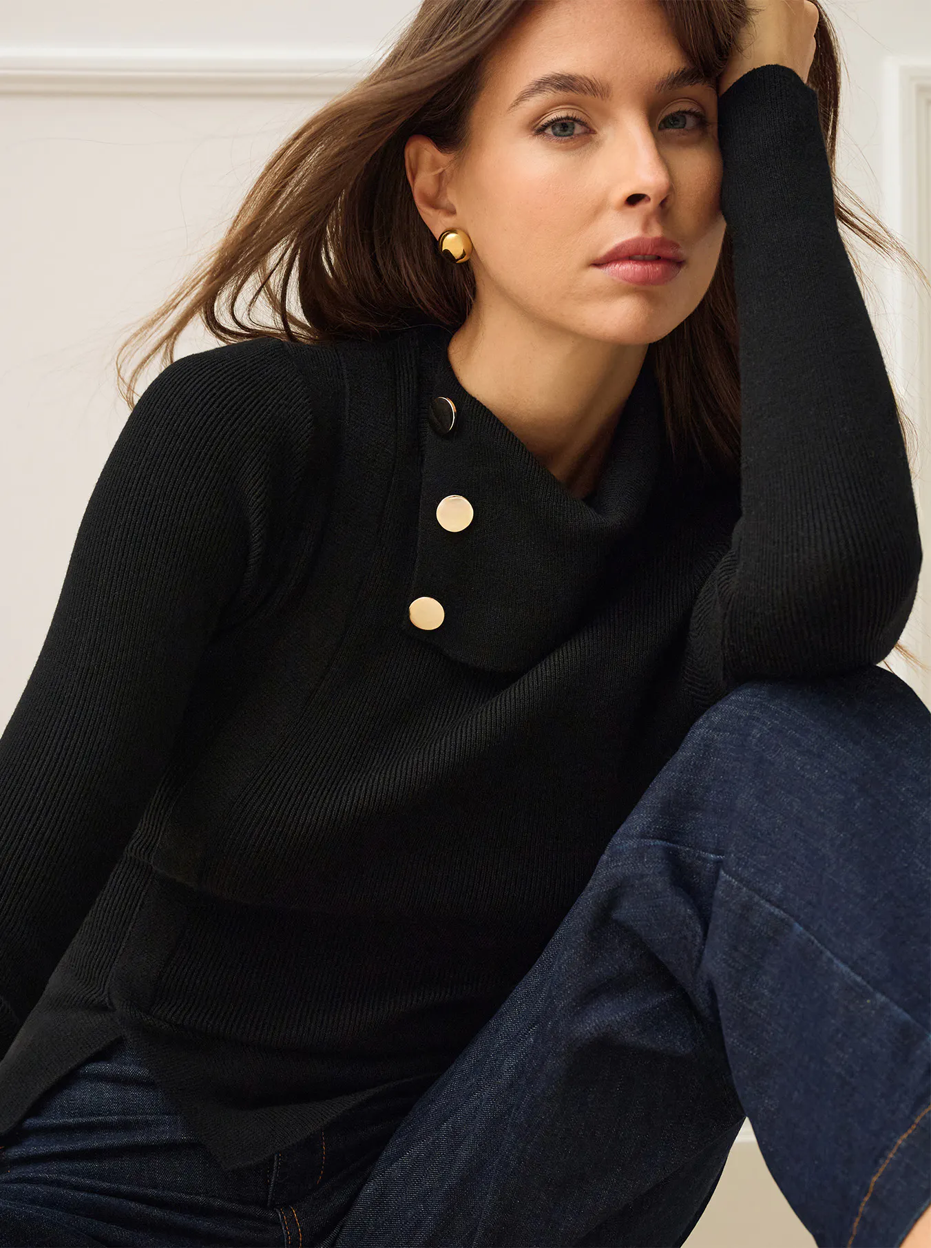 Fashion portrait of a woman in a black ribbed sweater with pearl buttons and dark jeans, leaning against a white chair in a bright room; poised, relaxed mood.
