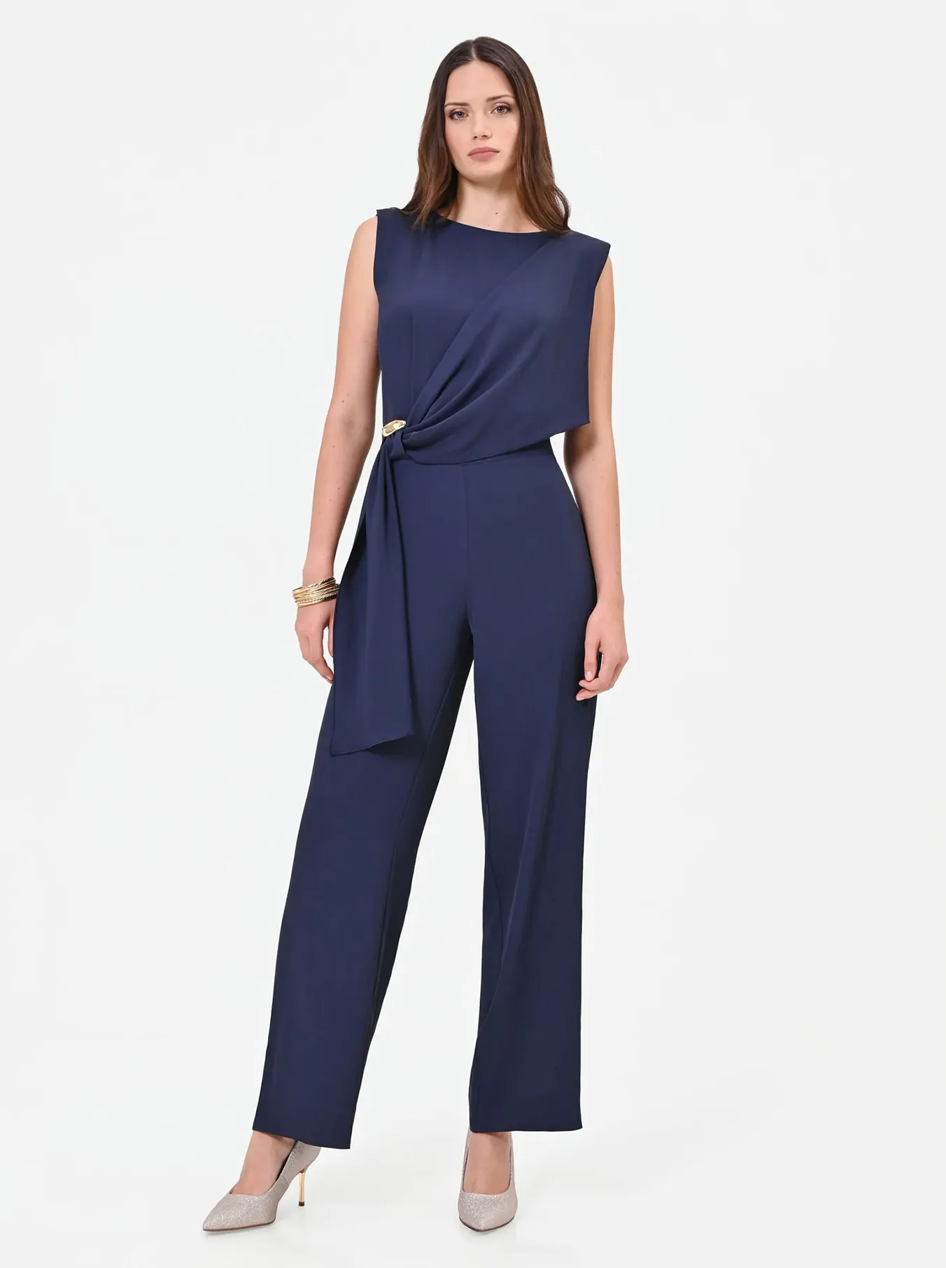 A woman stands confidently in a navy blue jumpsuit with a draped design and a knot detail. She wears beige heels and gold bracelets, exuding elegance and poise.