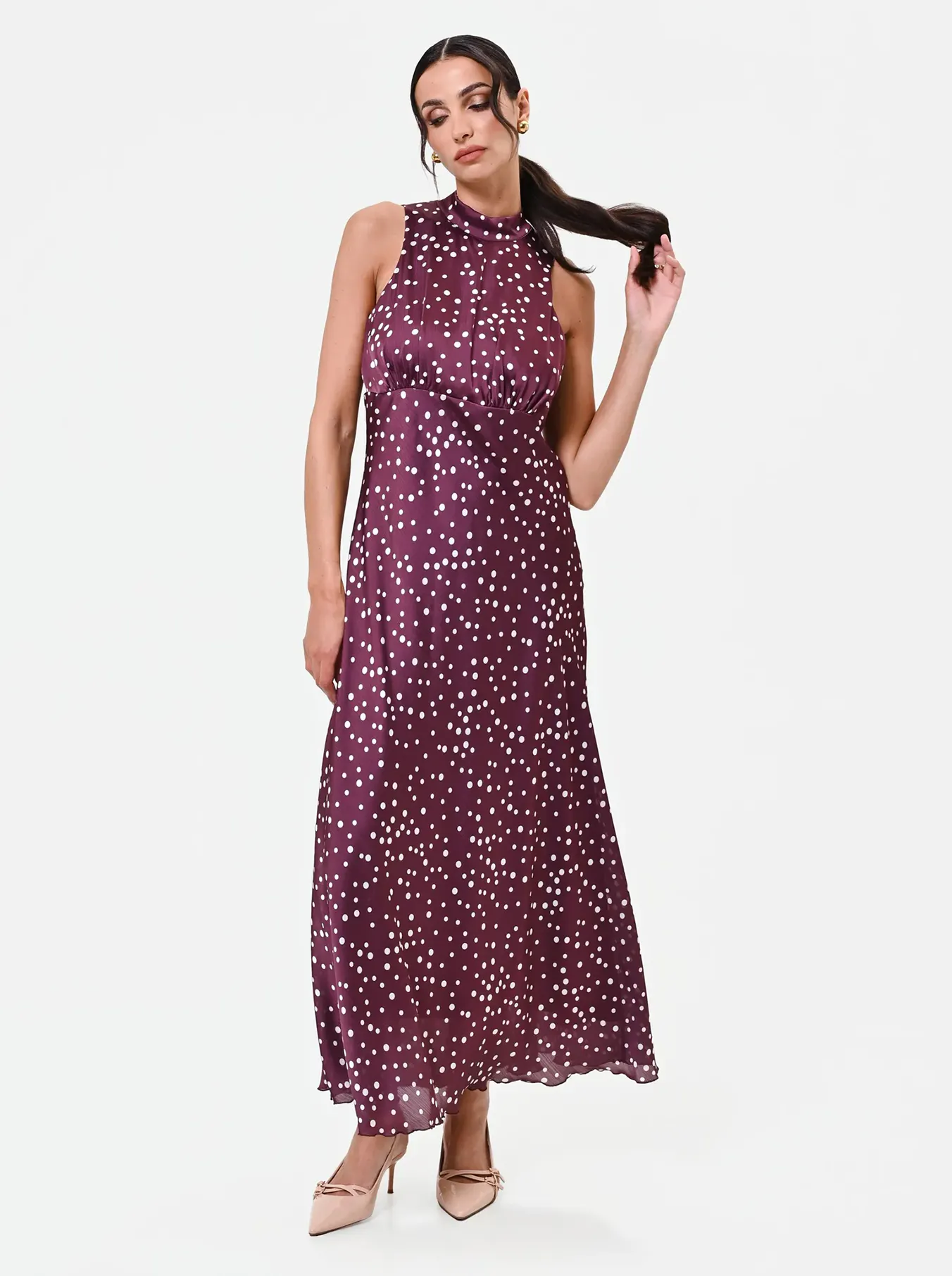 Woman in a sleeveless maroon polka-dot maxi dress, holding a ponytail, wears nude pointed heels. Elegant and poised against a white background.