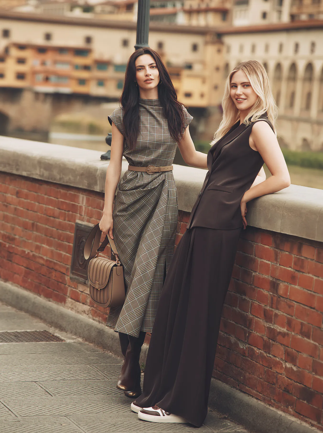 Two models pose outdoors on a brick bridge overlooking a scenic Italian river and cityscape. The brunette model on the left wears a brown plaid midi dress with a beige belt, carrying a brown leather handbag and wearing brown boots. The blonde model on the right leans against the wall in a dark brown sleeveless tailored vest and wide-leg trousers paired with stylish sneakers.