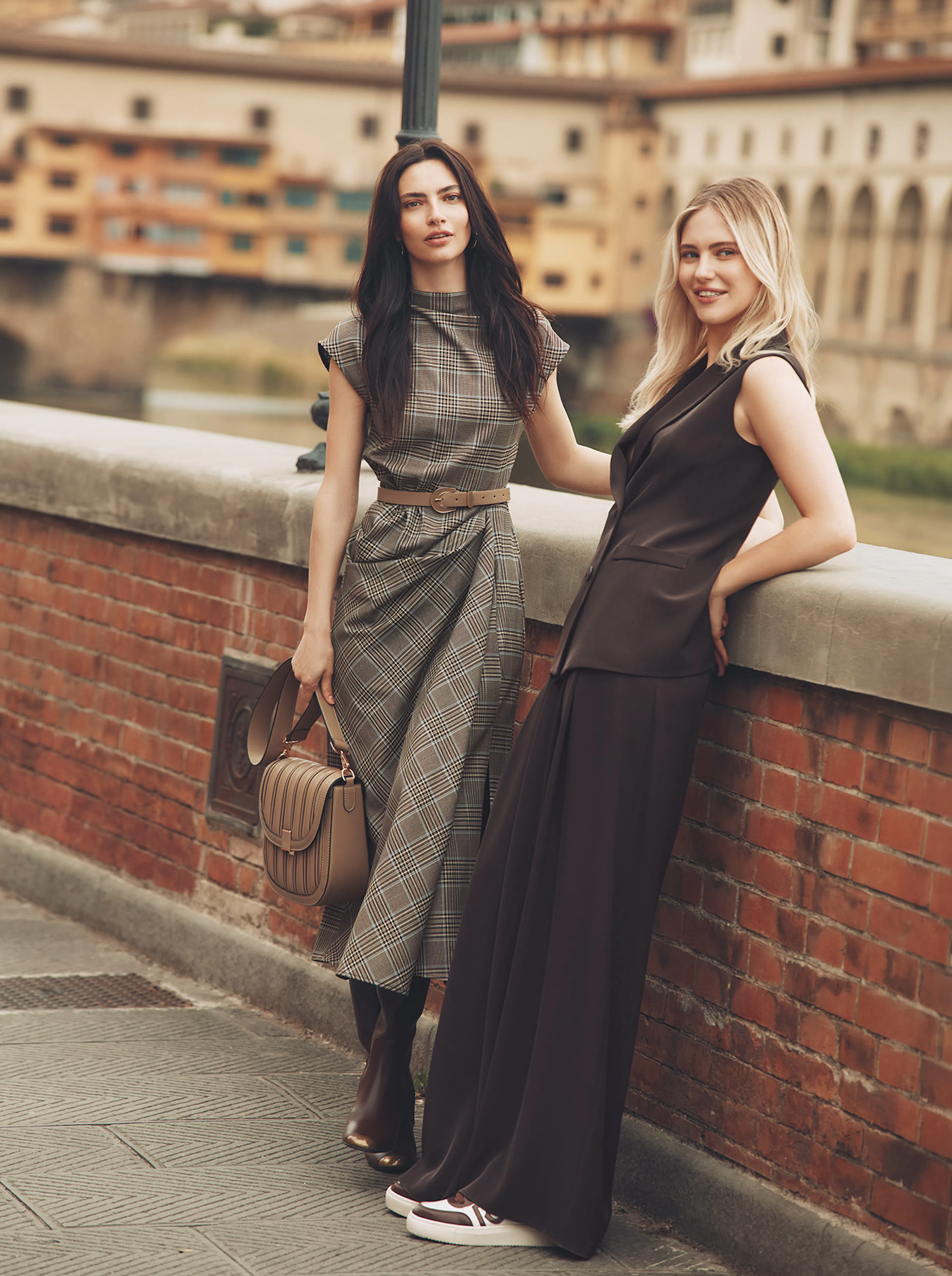 Two models pose outdoors on a brick bridge overlooking a scenic Italian river and cityscape. The brunette model on the left wears a brown plaid midi dress with a beige belt, carrying a brown leather handbag and wearing brown boots. The blonde model on the right leans against the wall in a dark brown sleeveless tailored vest and wide-leg trousers paired with stylish sneakers.