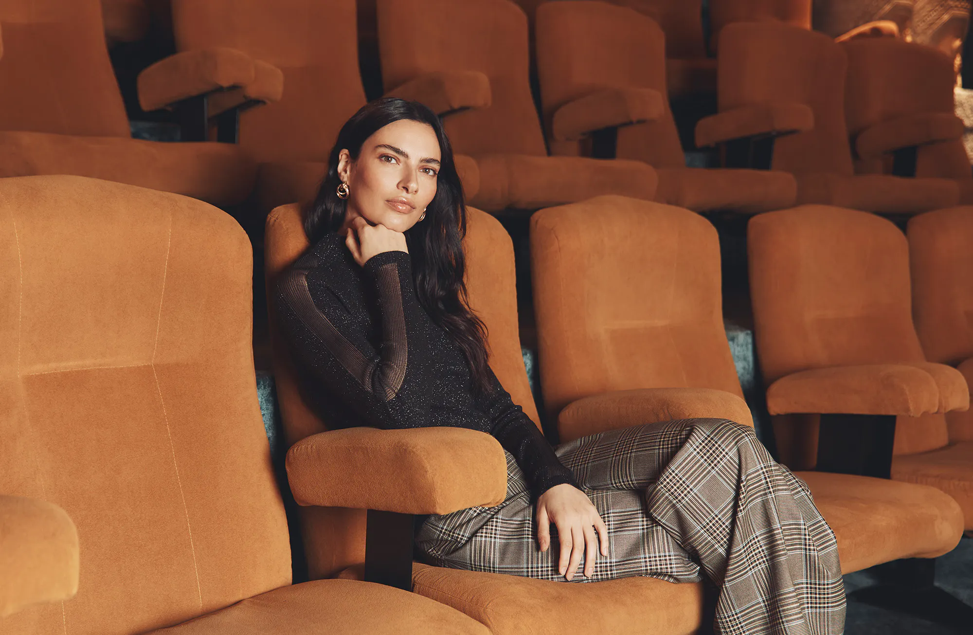A brunette model sits in an orange velvet seat inside a classic movie theater, resting her chin thoughtfully on her hand while looking at the camera. She is wearing a black long-sleeved top with sheer mesh panels and a subtle shimmer, paired with wide-leg plaid trousers.
