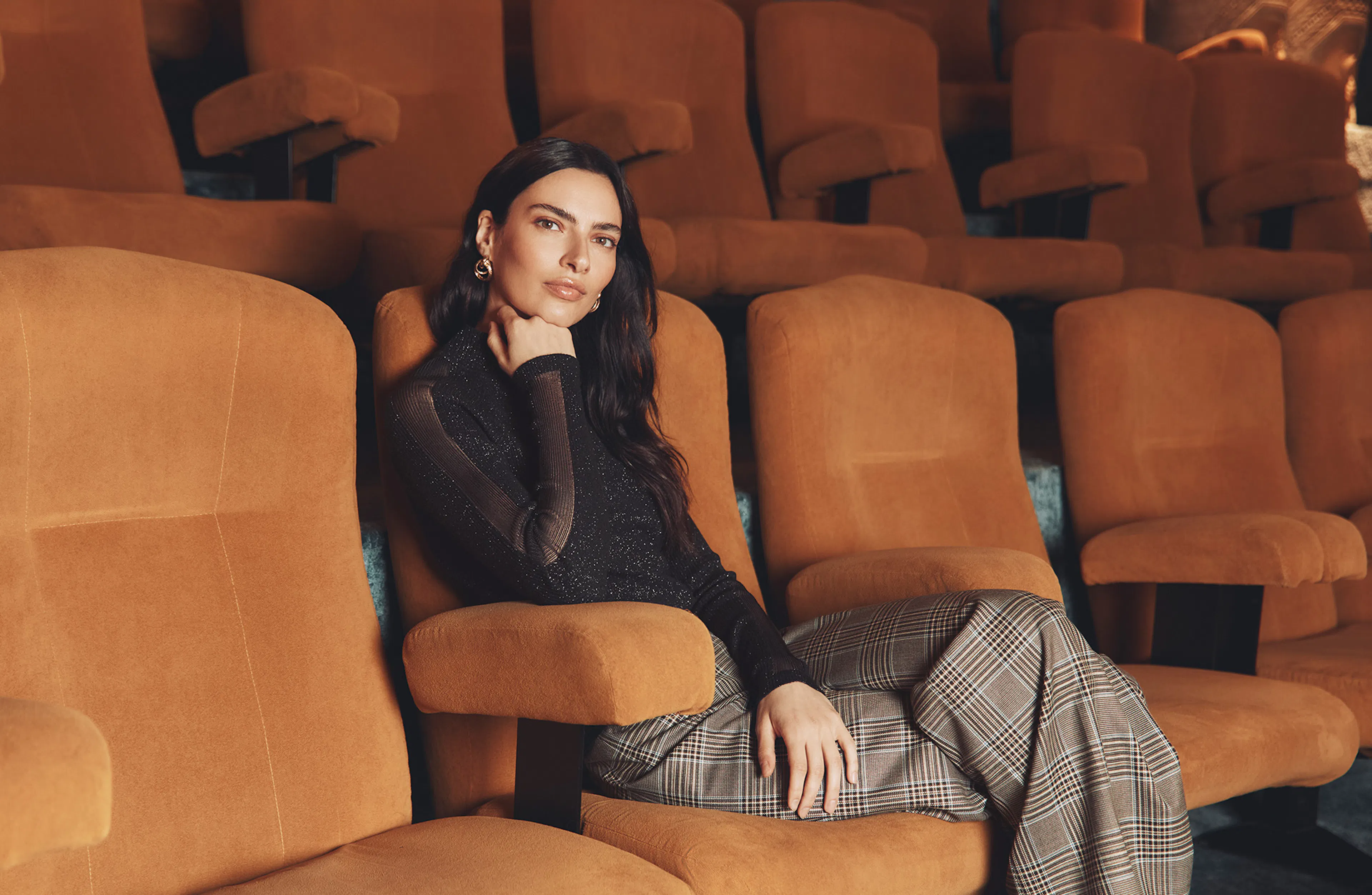 A brunette model sits in an orange velvet seat inside a classic movie theater, resting her chin thoughtfully on her hand while looking at the camera. She is wearing a black long-sleeved top with sheer mesh panels and a subtle shimmer, paired with wide-leg plaid trousers.