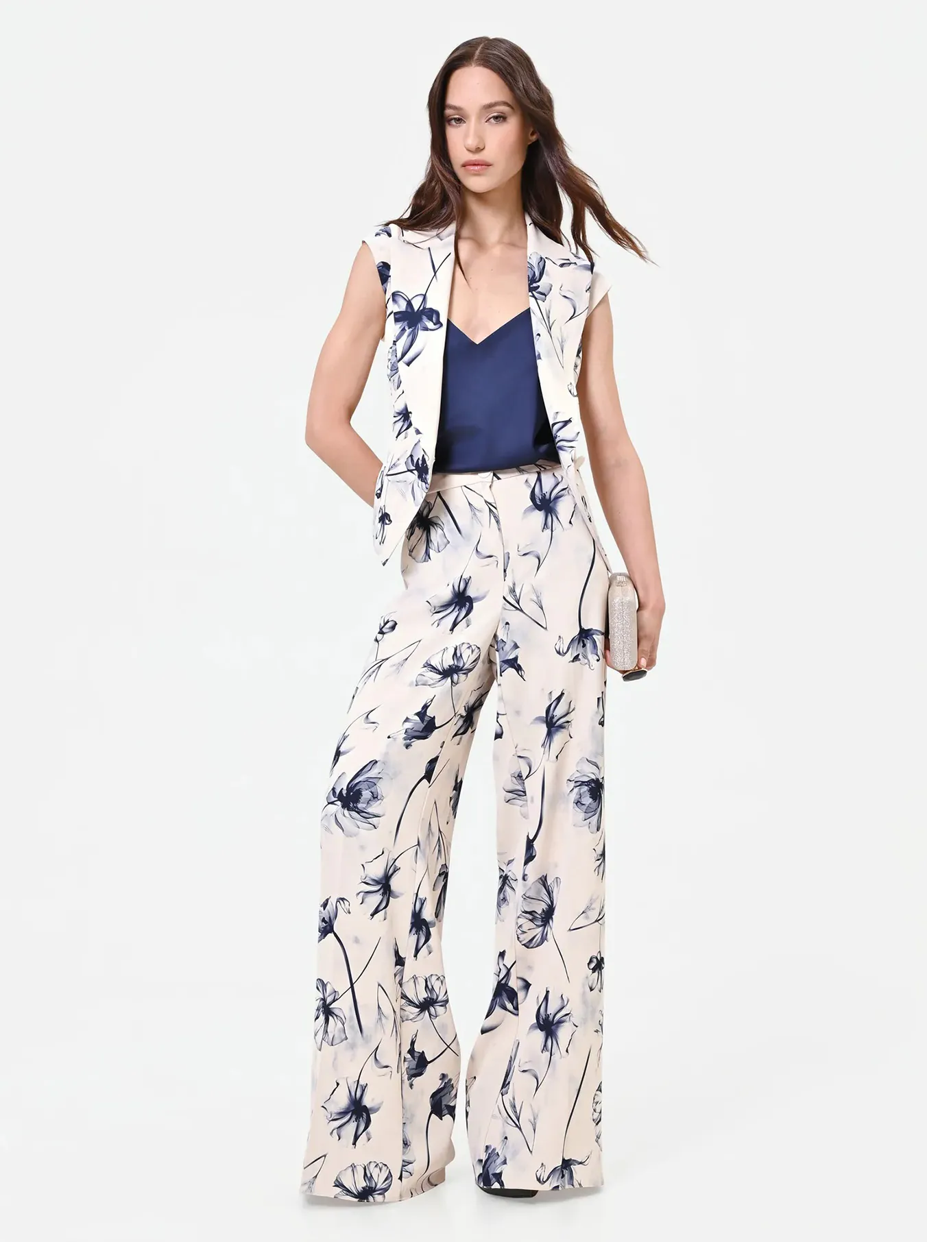 Woman in a sleeveless floral suit with wide-leg pants and a navy blouse, holding a clutch. She exudes a chic and confident vibe against a plain background.