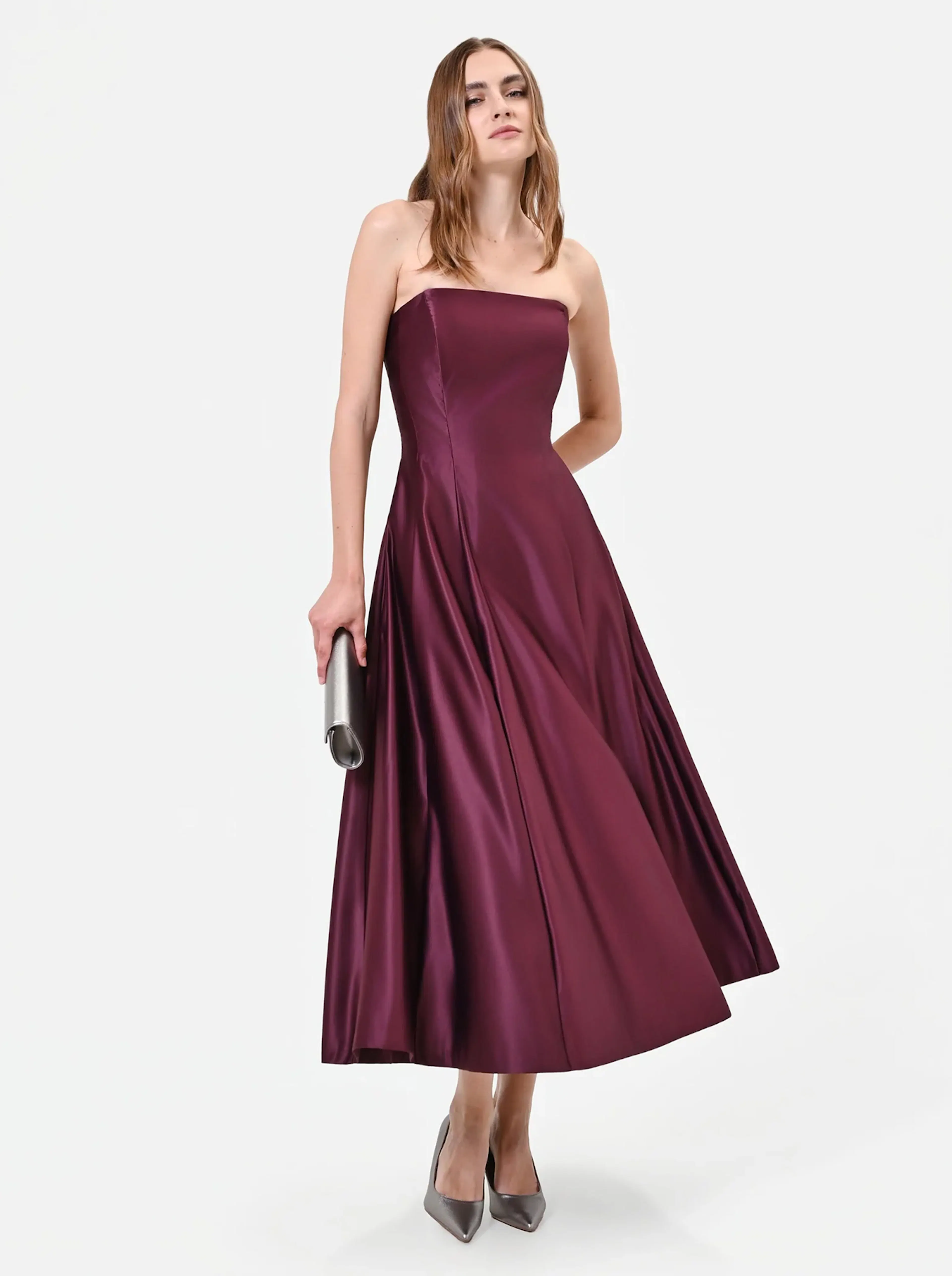 A woman in a strapless, burgundy satin gown holds a silver clutch. She stands confidently in gray heels against a plain white background.