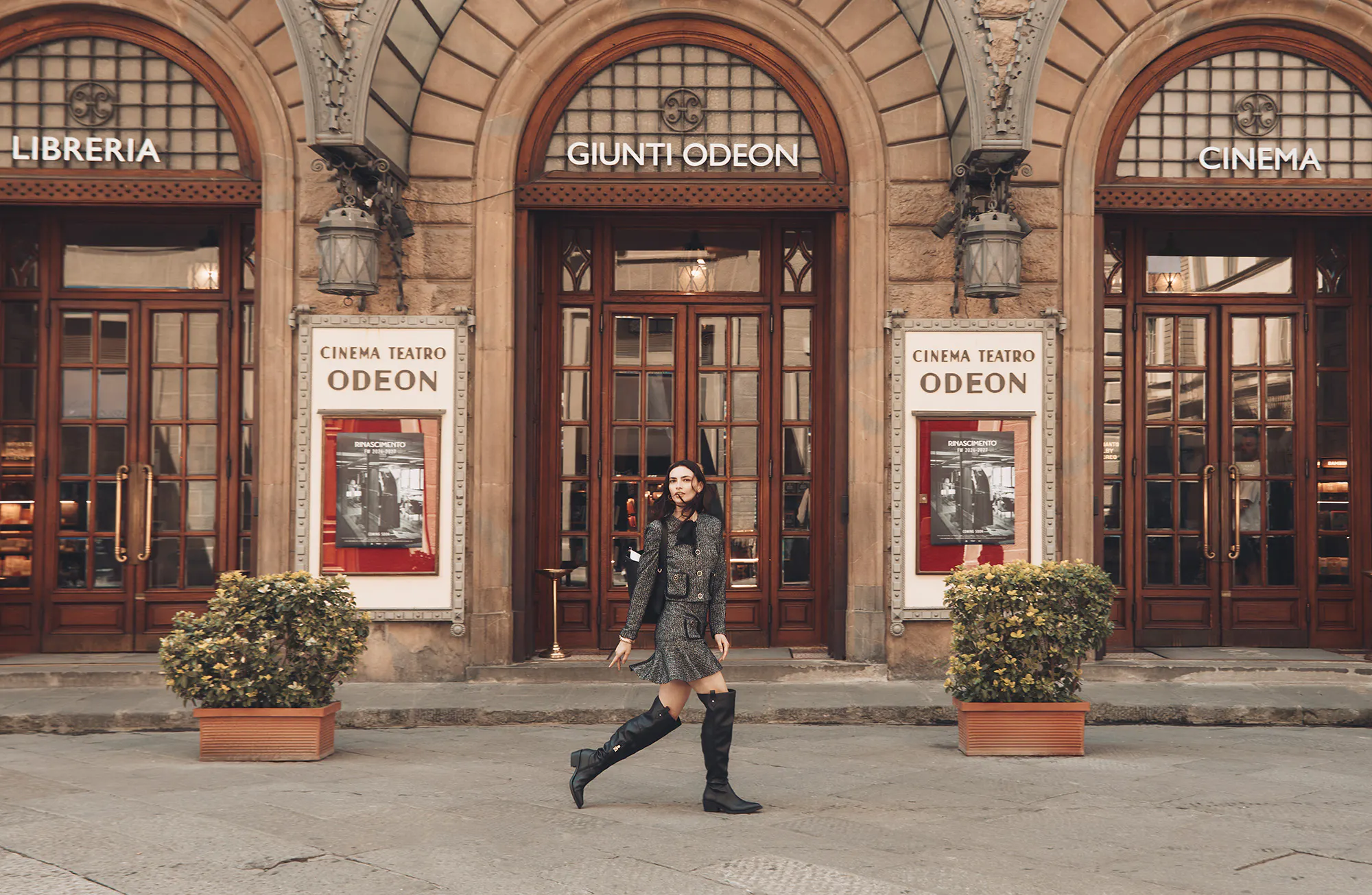 A wide shot showing a woman walking past the historic entrance of the Giunti Odeon cinema and library in Florence, Italy, which features arched doorways and large poster displays. The woman is wearing a grey tweed skirt suit and black knee-high boots and looks towards the viewer. Potted shrubs are placed in front of the building.