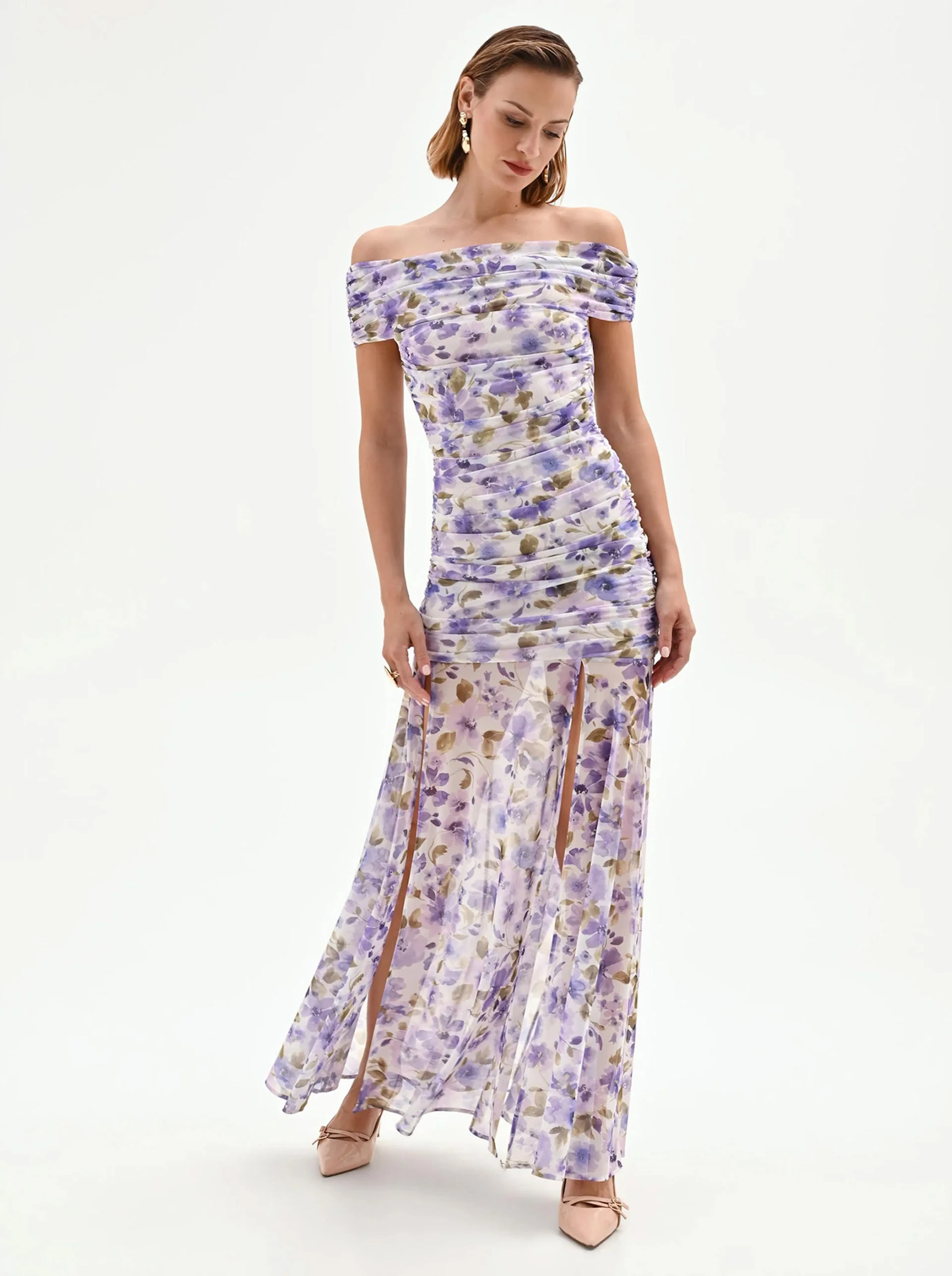 Woman in an off-shoulder, floral-print gown with purple and beige hues stands gracefully on a white background, exuding elegance and style.