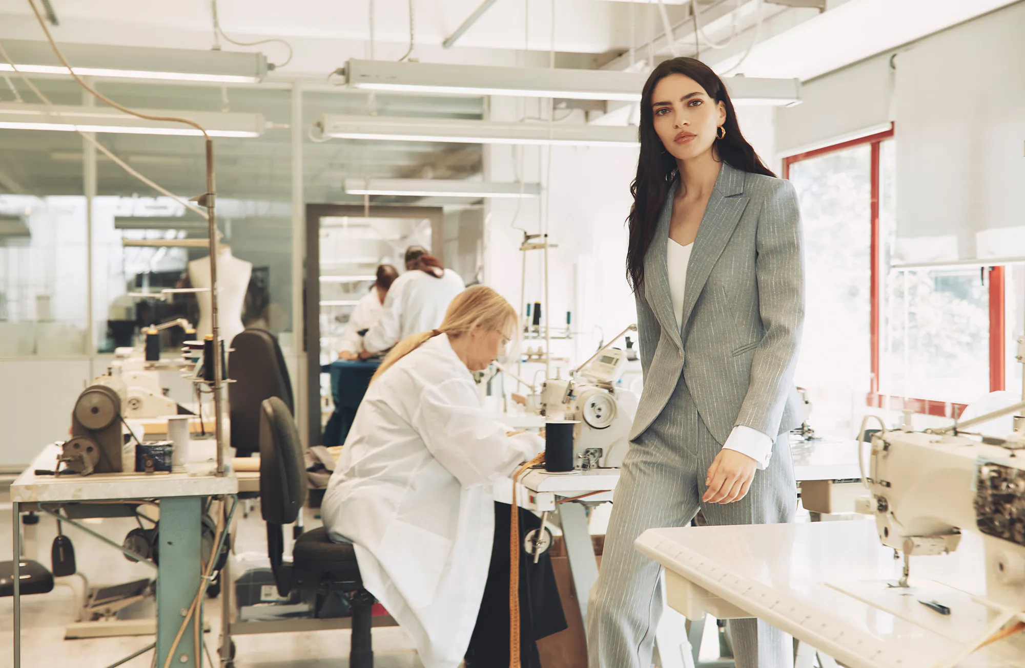 Woman in a pinstripe suit standing in a bright sewing workshop with workers using sewing machines in the background.