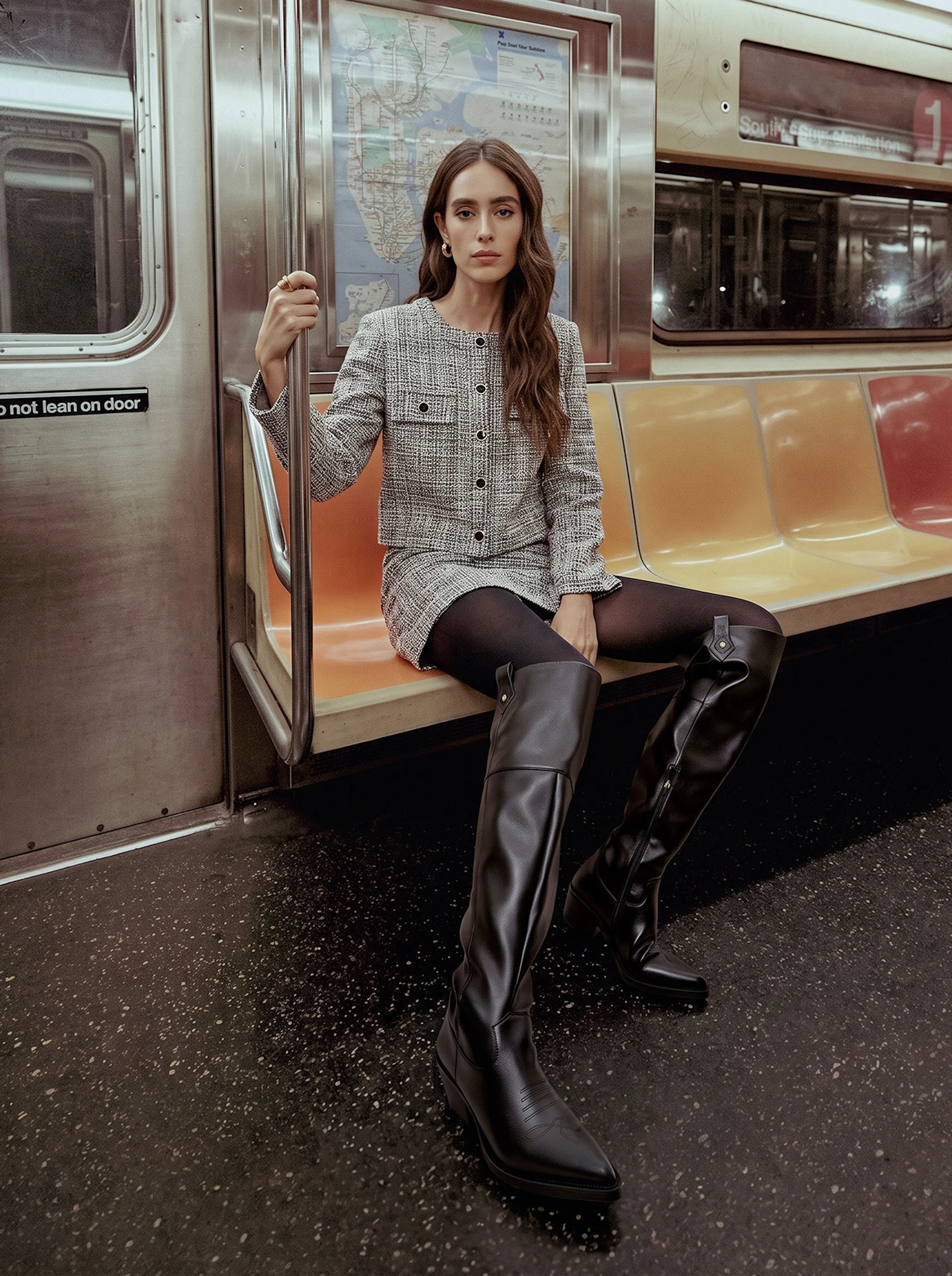 Young woman in a gray tweed outfit and black thigh-high boots sits on an orange subway seat, holding a pole with a calm, stylish expression.