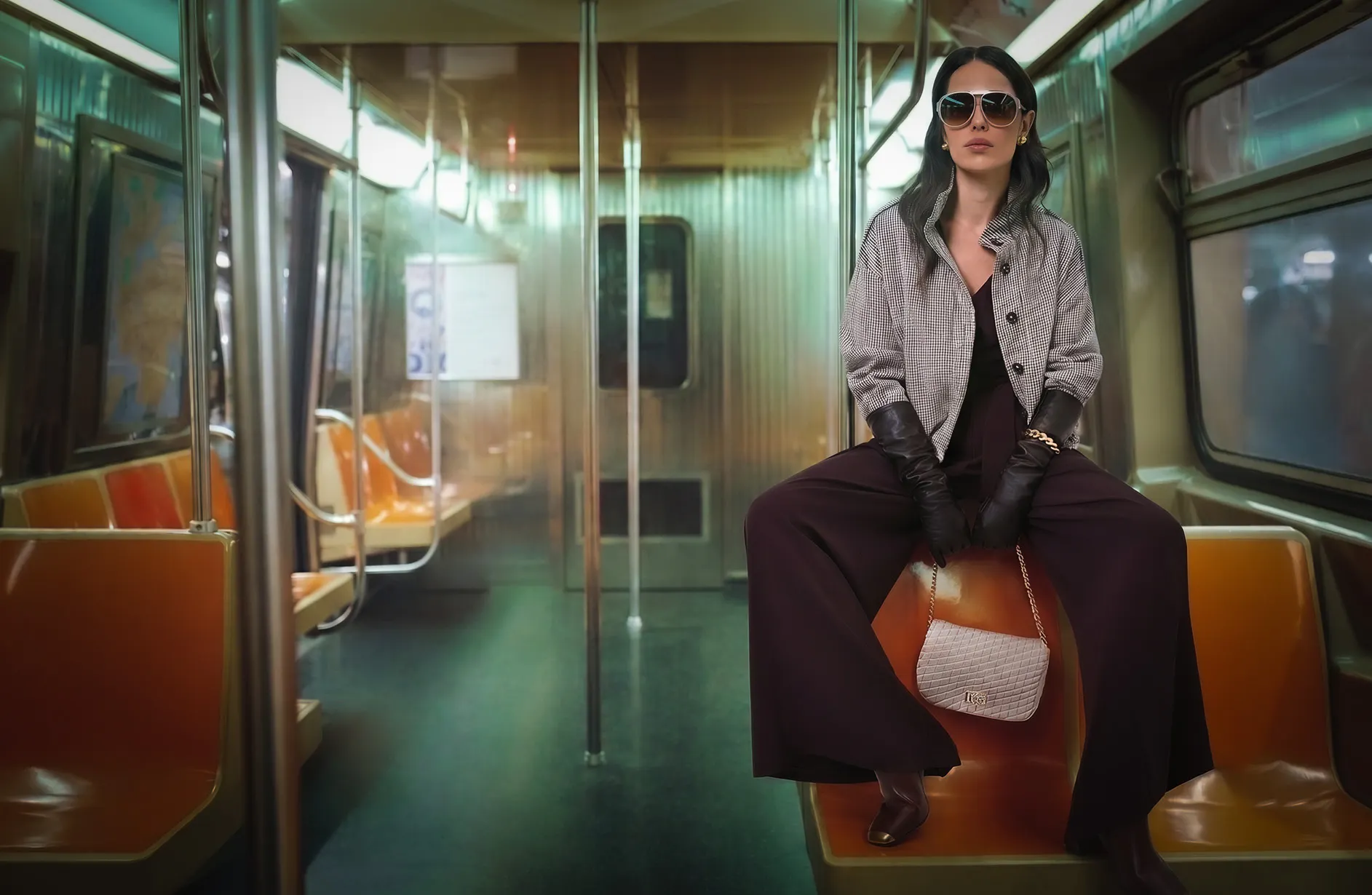 Woman in sunglasses sits confidently on an empty subway train, wearing a patterned jacket, gloves, and wide-leg trousers.