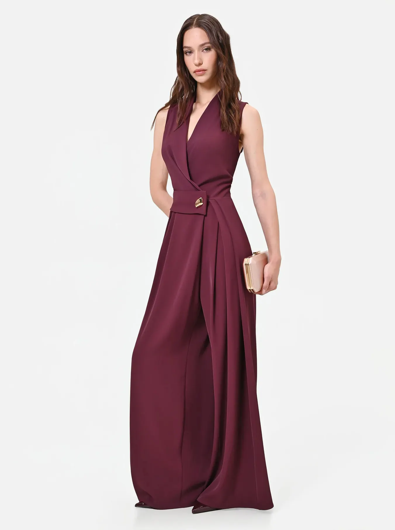 Woman in a sleeveless, deep maroon jumpsuit holding a gold clutch. The elegant outfit features draped pants, conveying a sophisticated, stylish tone.