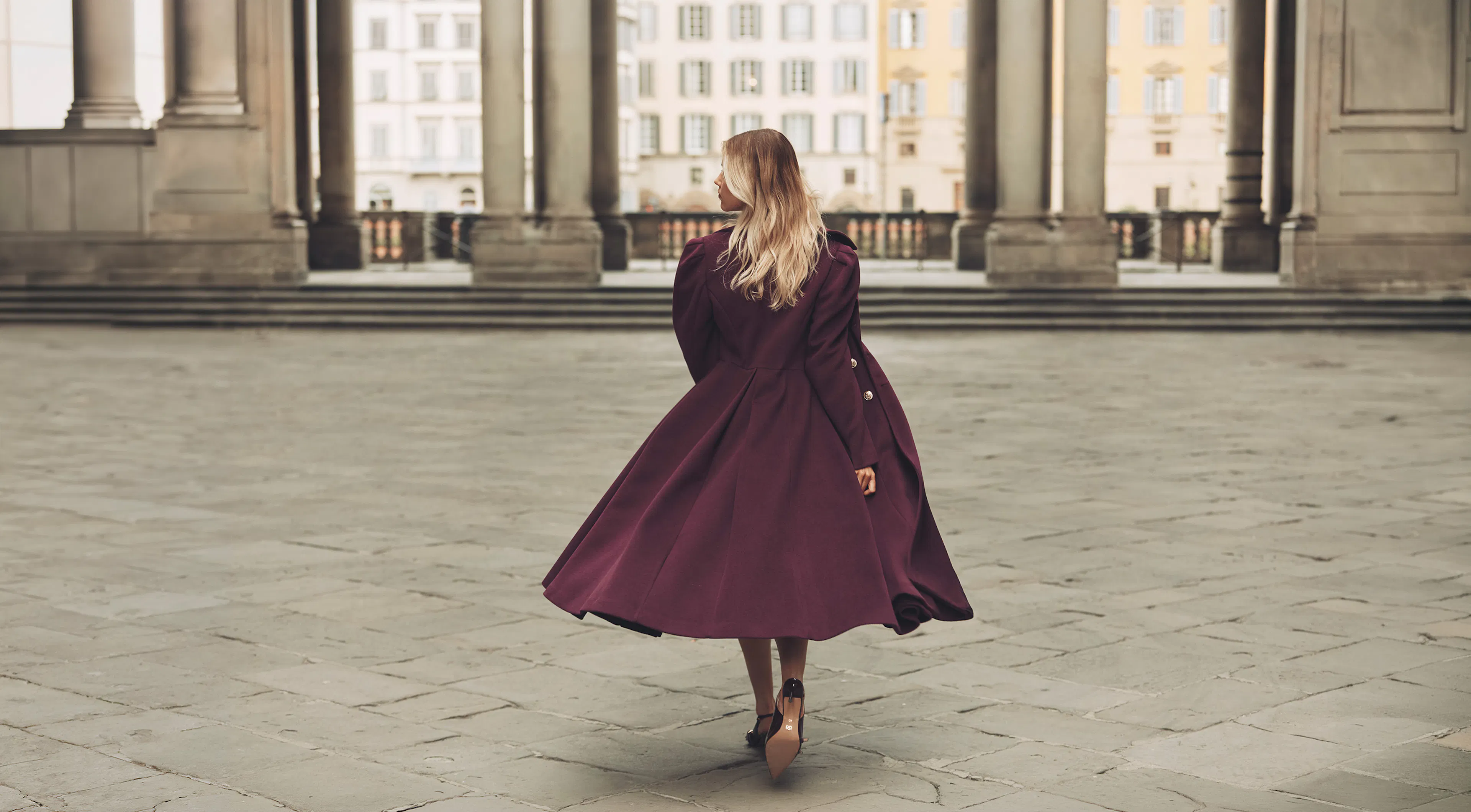 Woman in a flowing dark purple coat walking on a stone plaza with large columns and buildings in the background.
